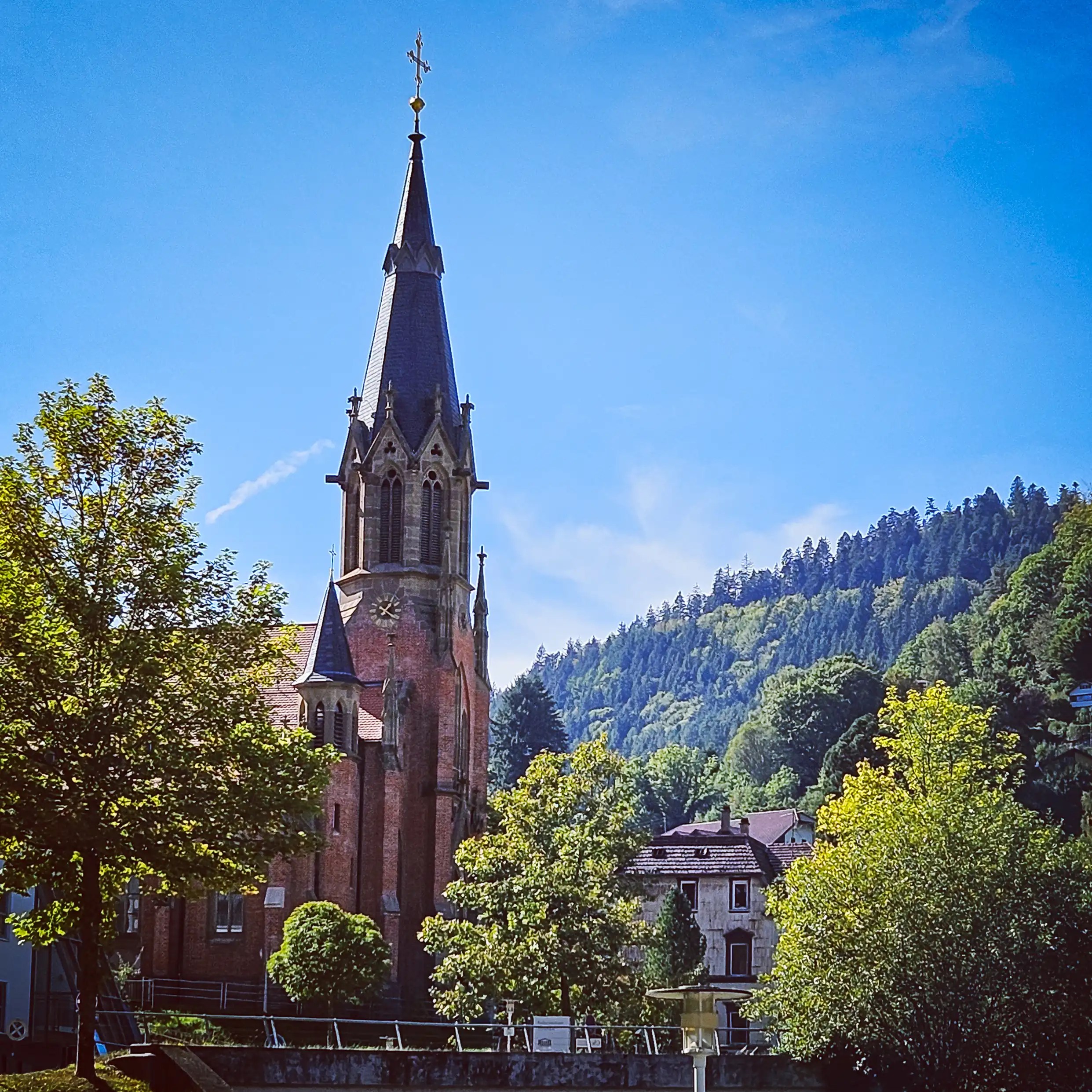 A tall church steeple rises above trees and rooftops in Bad Wildbad with forested hills behind it.