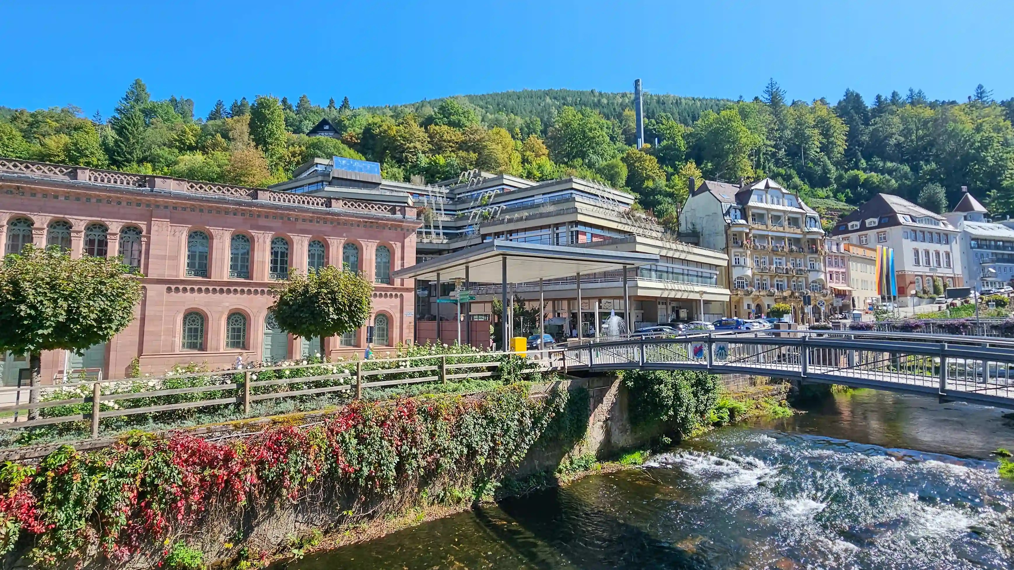 Buildings line the Enz River in Bad Wildbad, including the pink sandstone façade of Palais Thermal, with forested hills rising behind the town.
