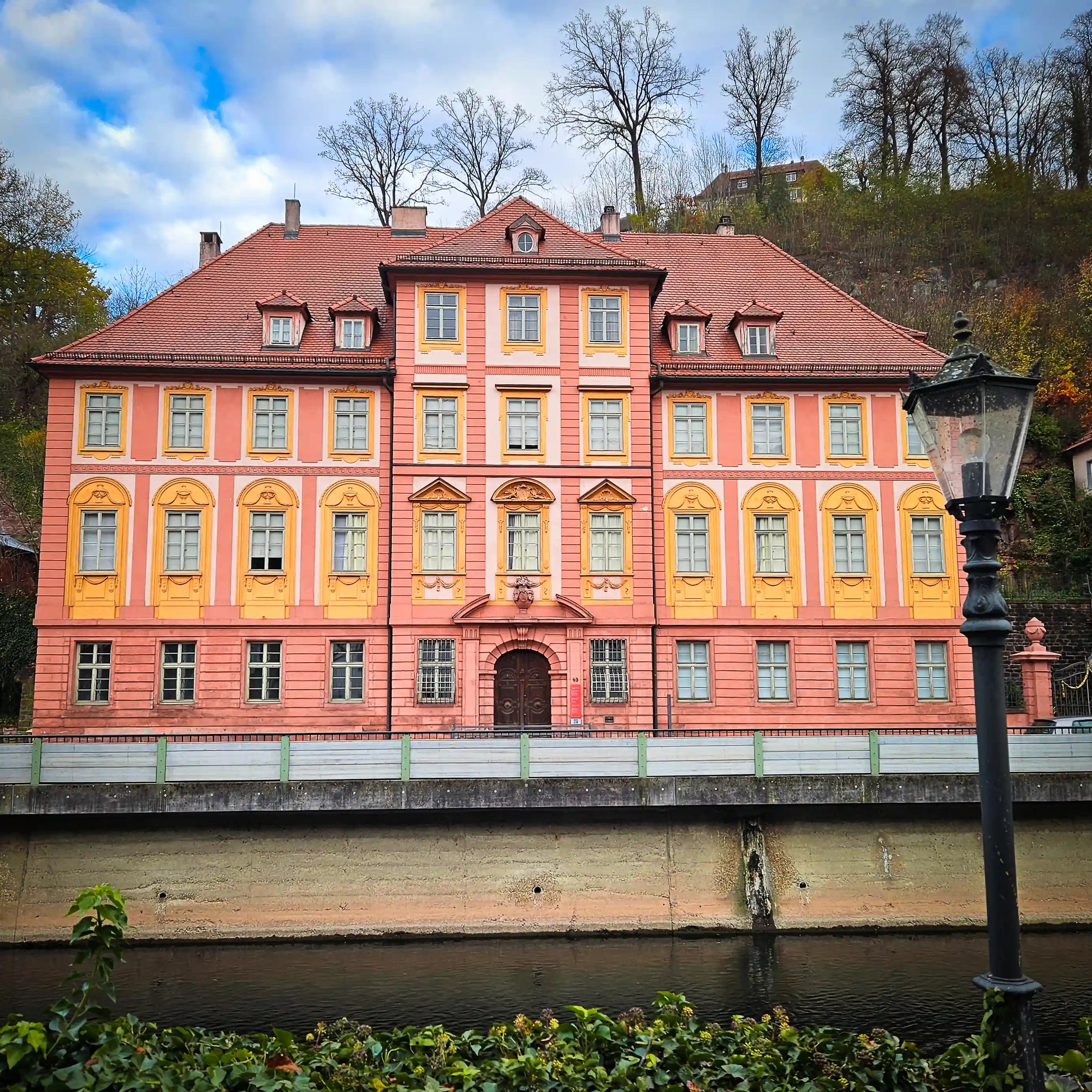 A large pink and yellow Baroque-style building stands along the river in Calw, with bare trees rising on the hillside behind it.