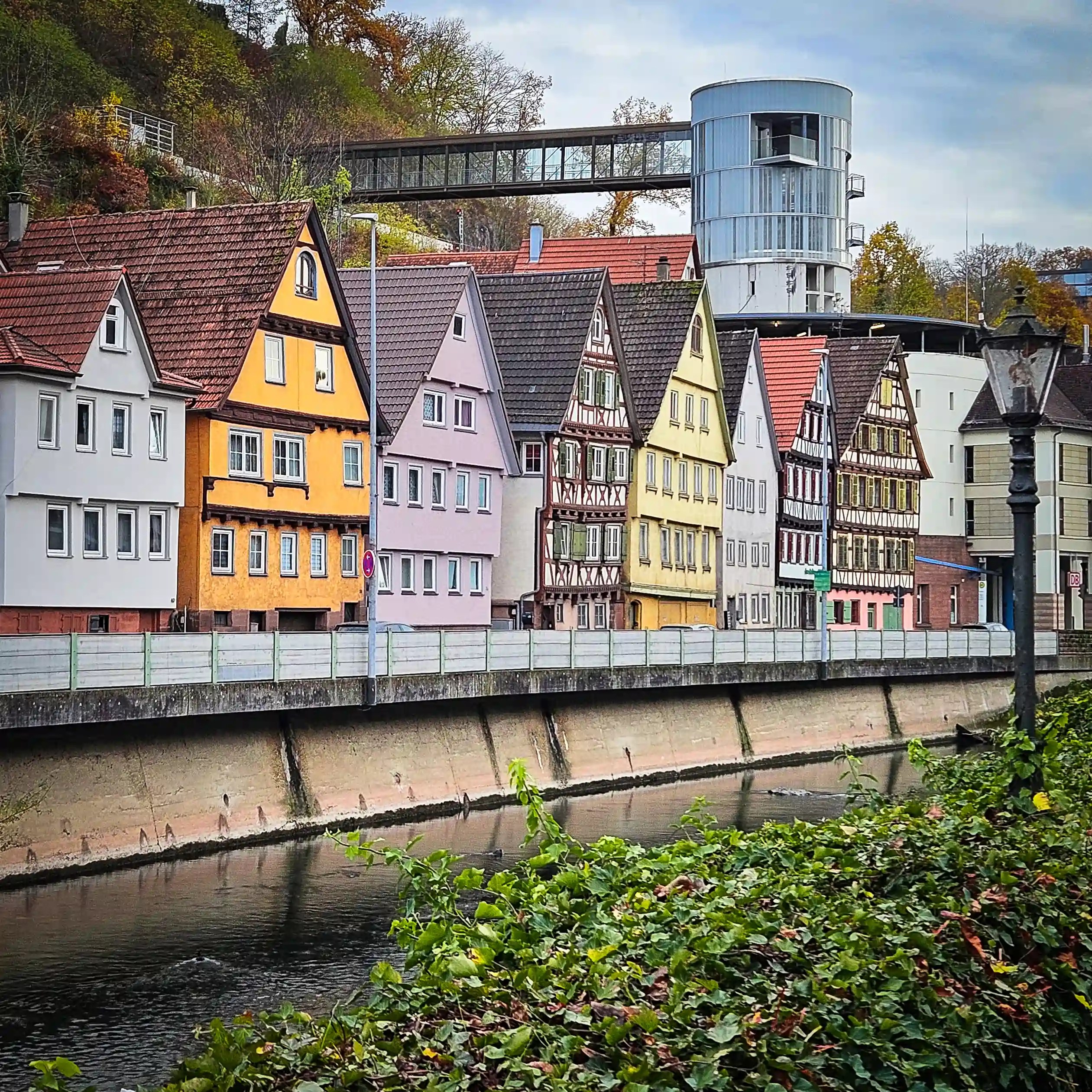 A row of colorful houses and half-timbered buildings lines the river in Calw, with a modern cylindrical glass tower and elevated walkway behind them.