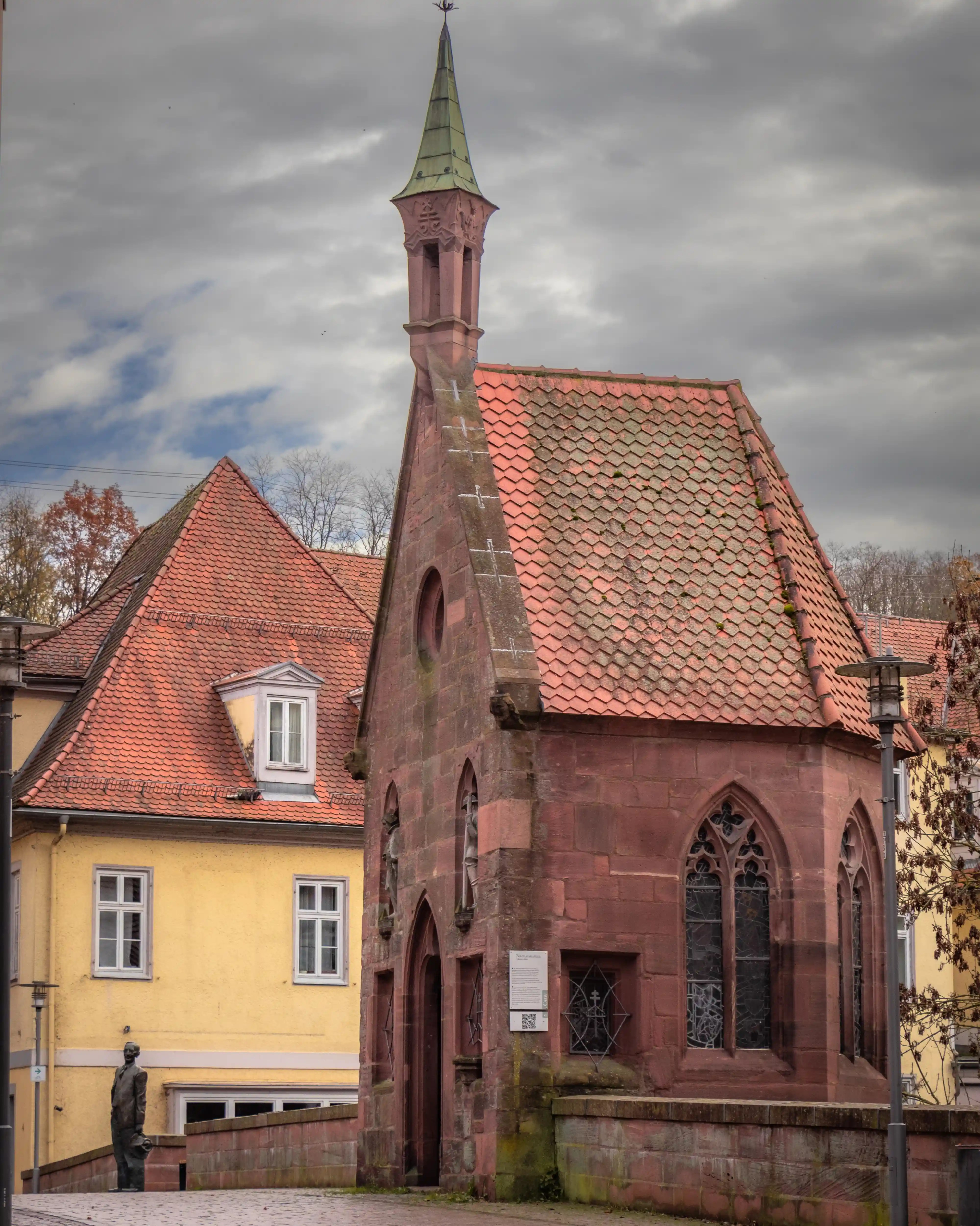A small red-sandstone chapel with a steep tiled roof and pointed tower stands beside yellow historic buildings in Calw, Germany.