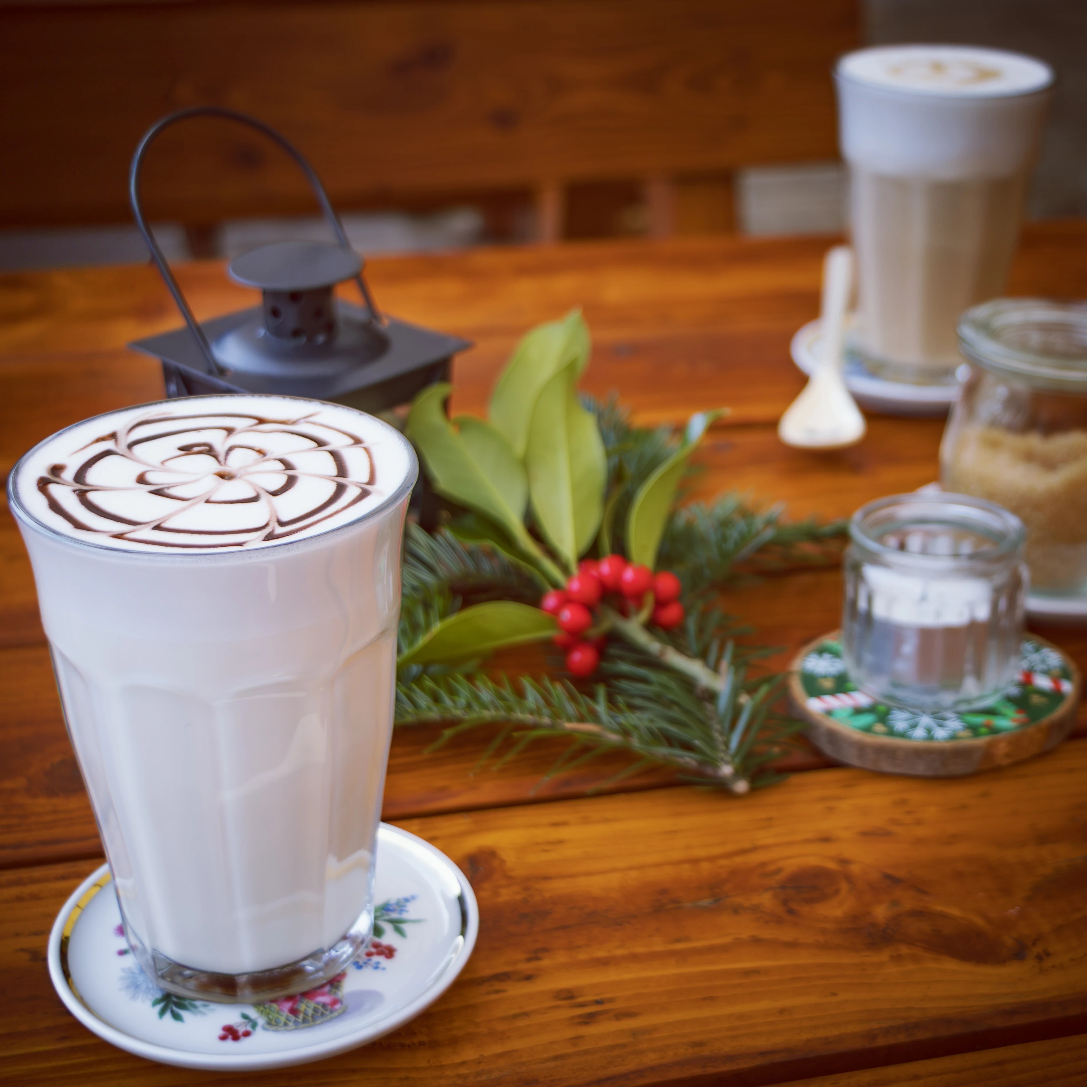 A glass of latte with a chocolate pattern sits on a wooden table decorated with evergreen and red berries, with another drink blurred in the background.
