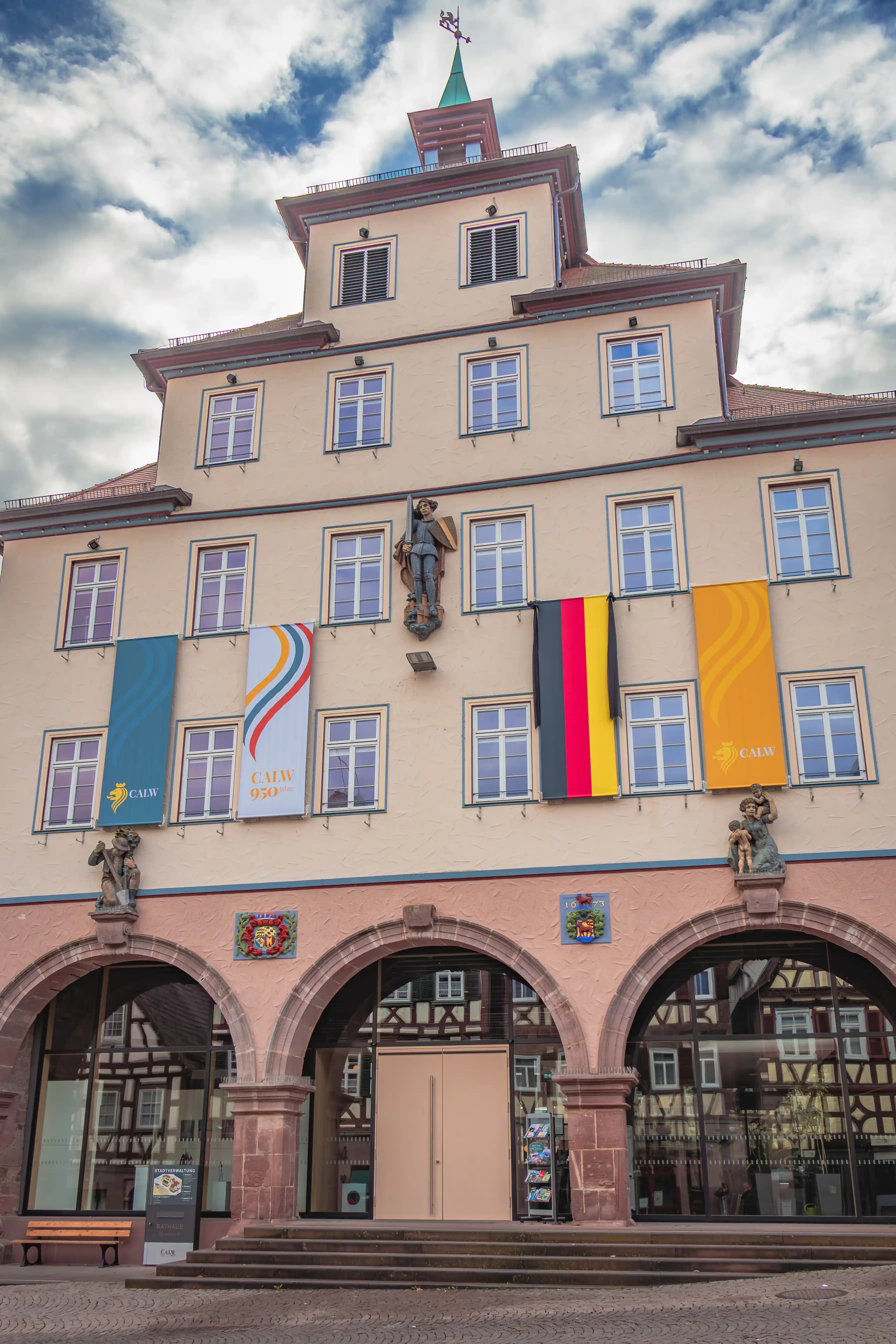 The façade of Calw’s Rathaus displays banners, statues, and arched entrances beneath a tower with a green spire.