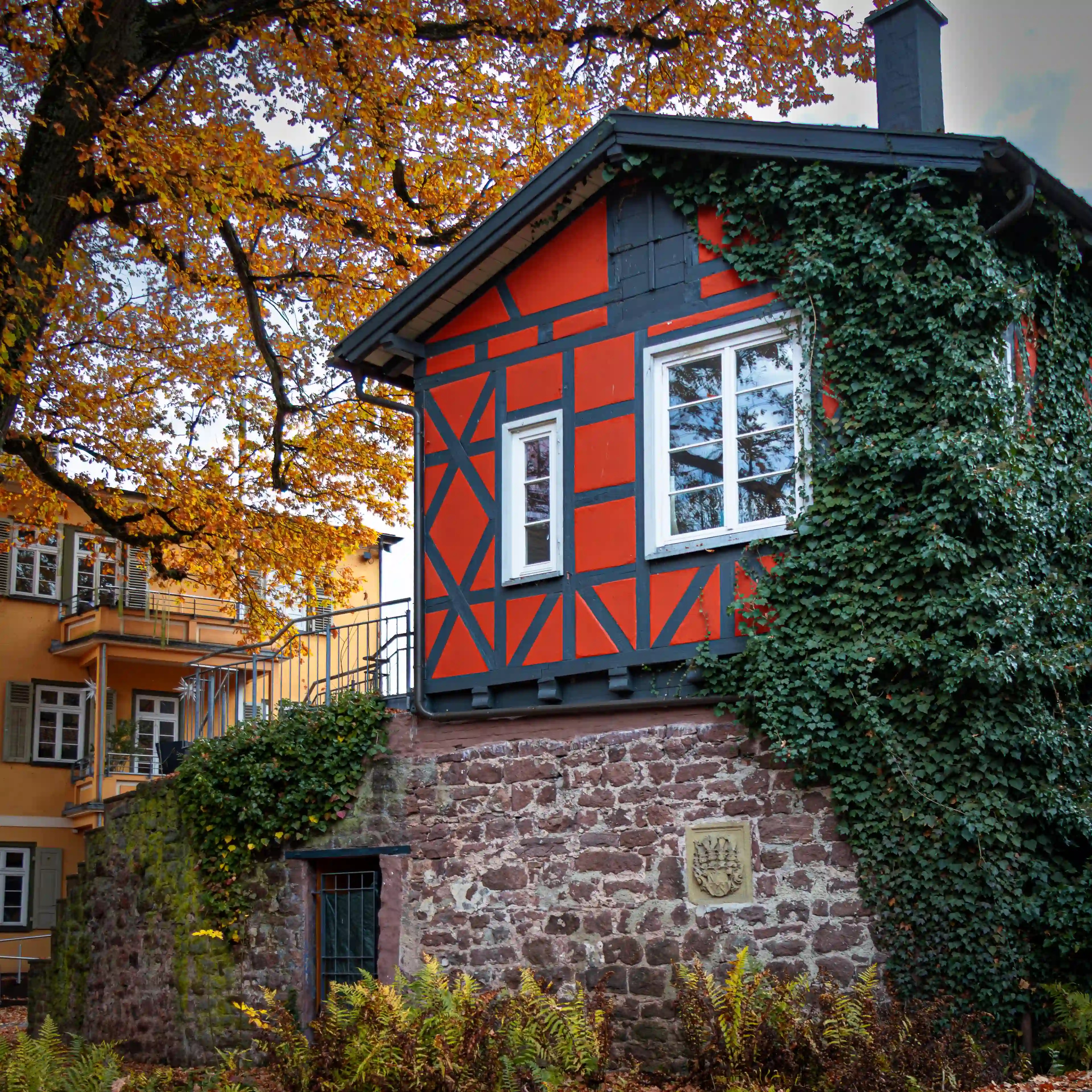 A small red-and-black half-timbered building stands on a stone base covered partly in ivy, surrounded by autumn foliage.