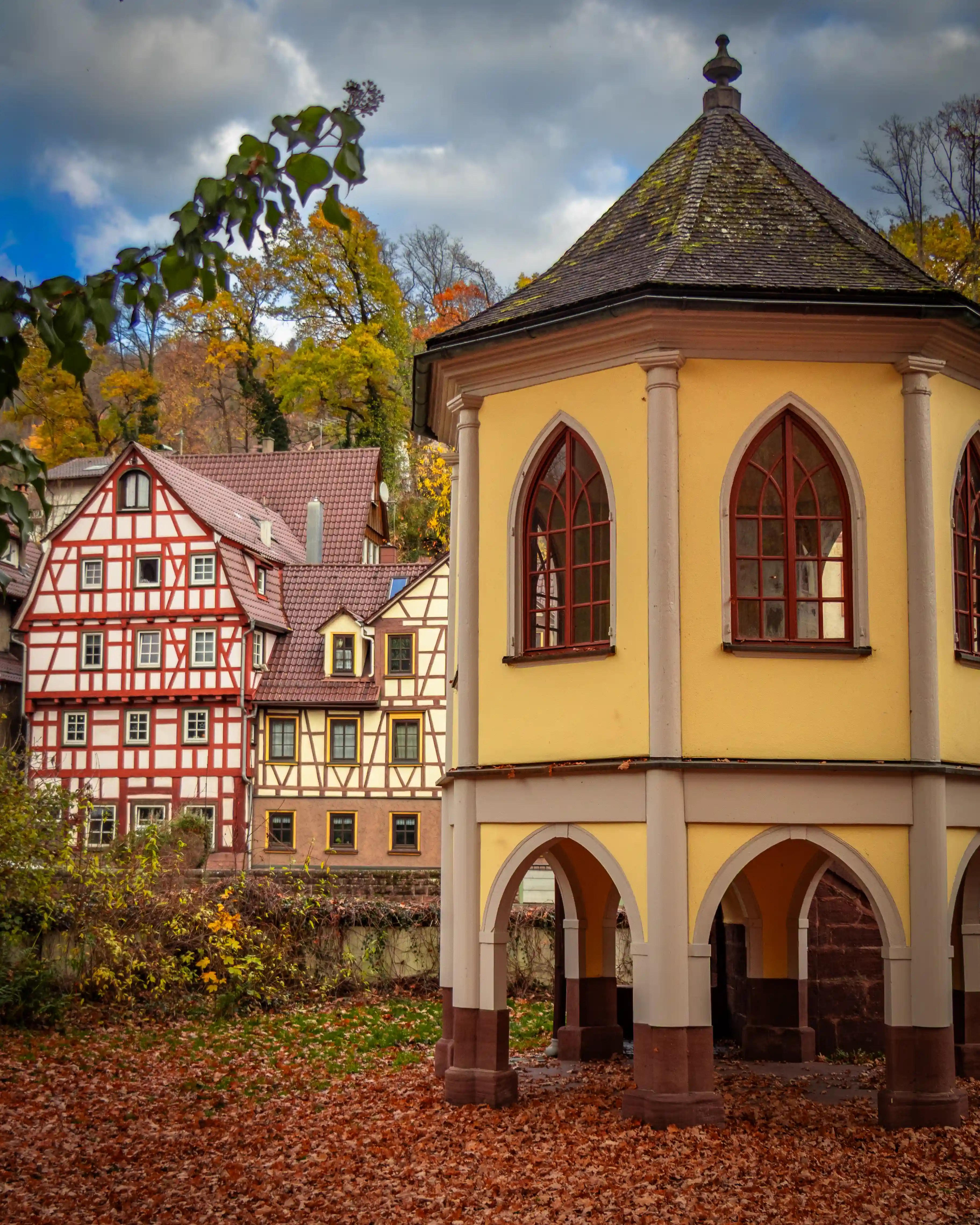 A yellow octagonal pavilion with arched openings stands in front of several half-timbered houses and autumn trees in Calw.