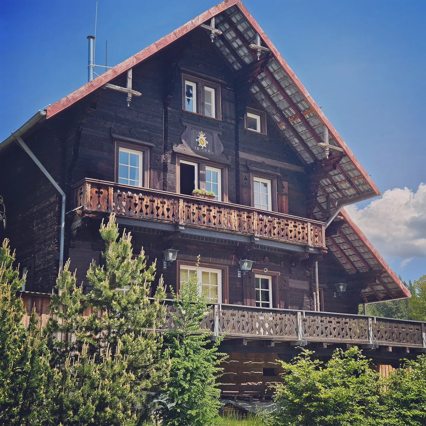A dark wooden Black Forest house with balconies stands among trees under a blue sky.