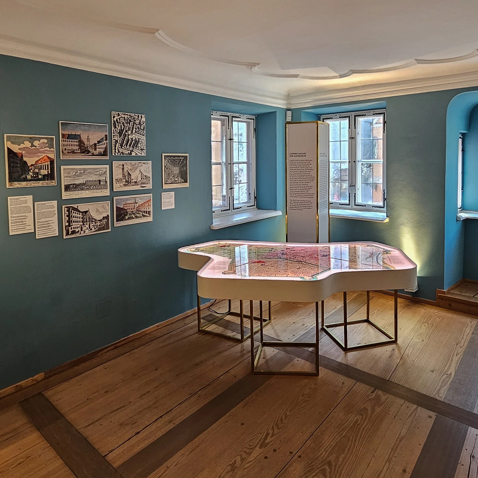 Interior exhibit room of the Leopold Mozart House in Augsburg with historic prints, windows, and an illuminated city map table.