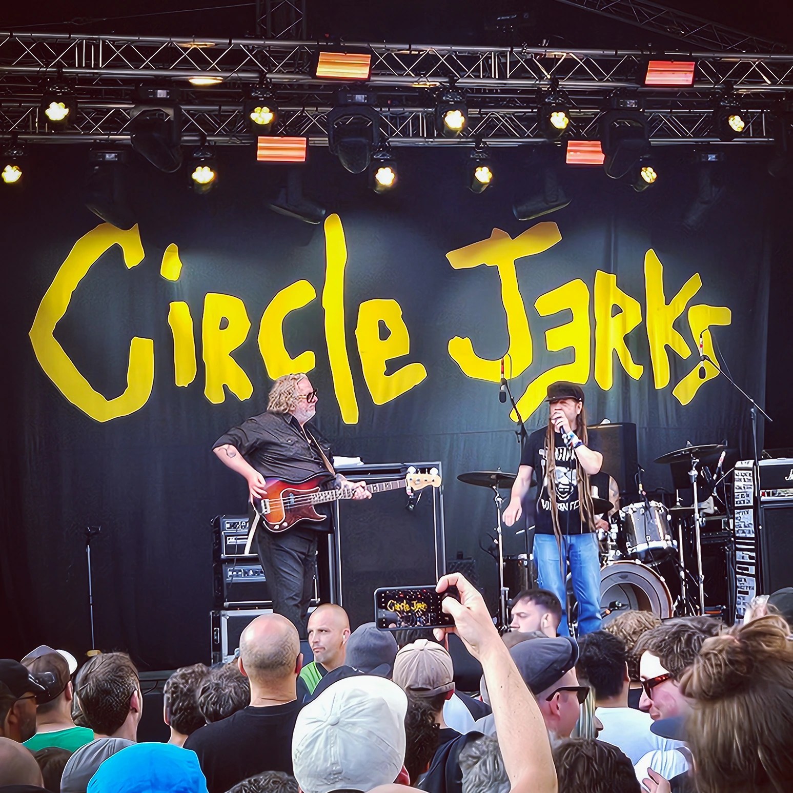 The band Circle Jerks performing on stage at an outdoor concert in Augsburg with a crowd gathered close to the stage.