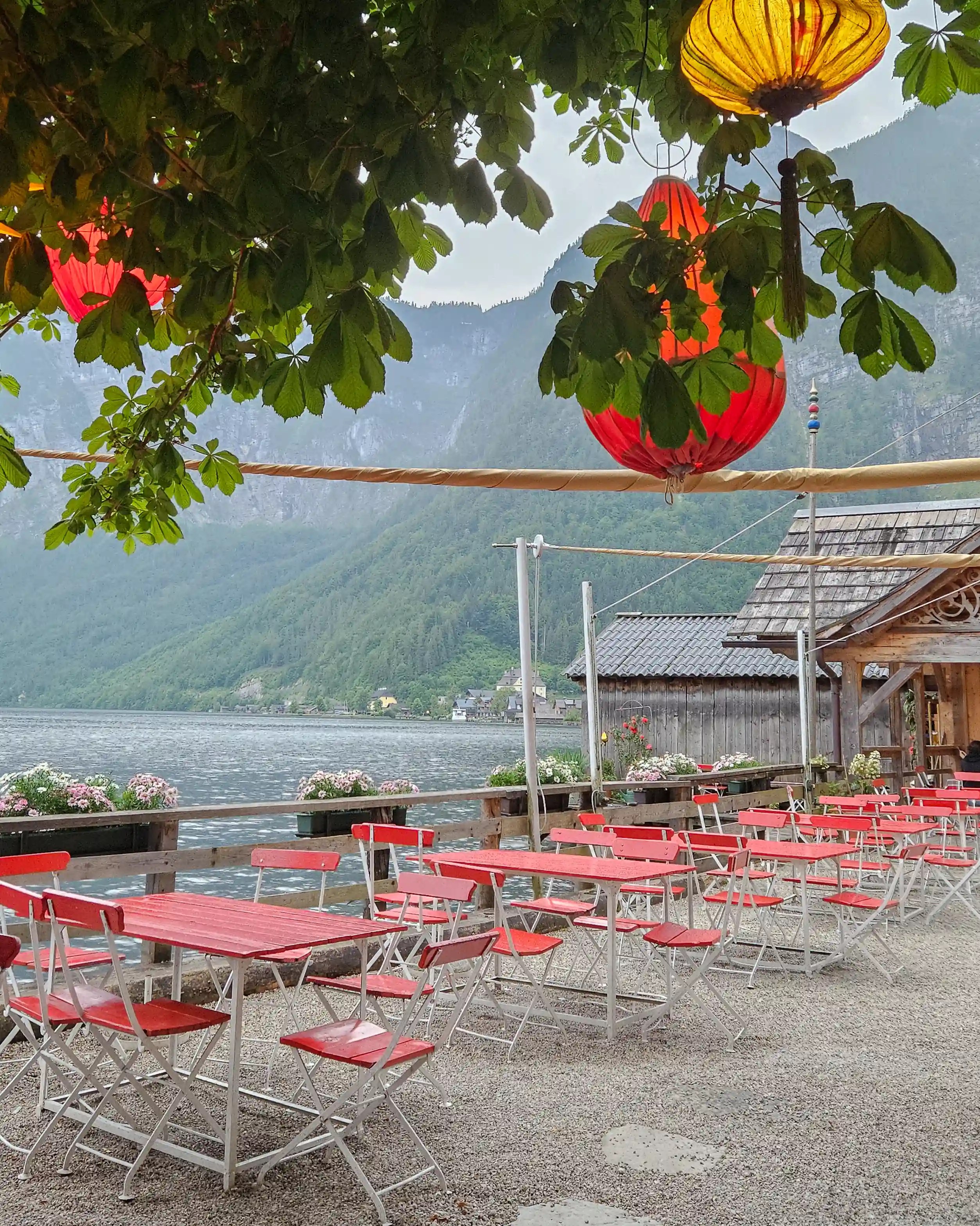 Red café tables sit beside the lake in Hallstatt beneath hanging lanterns and leafy branches, with mountains rising in the background.