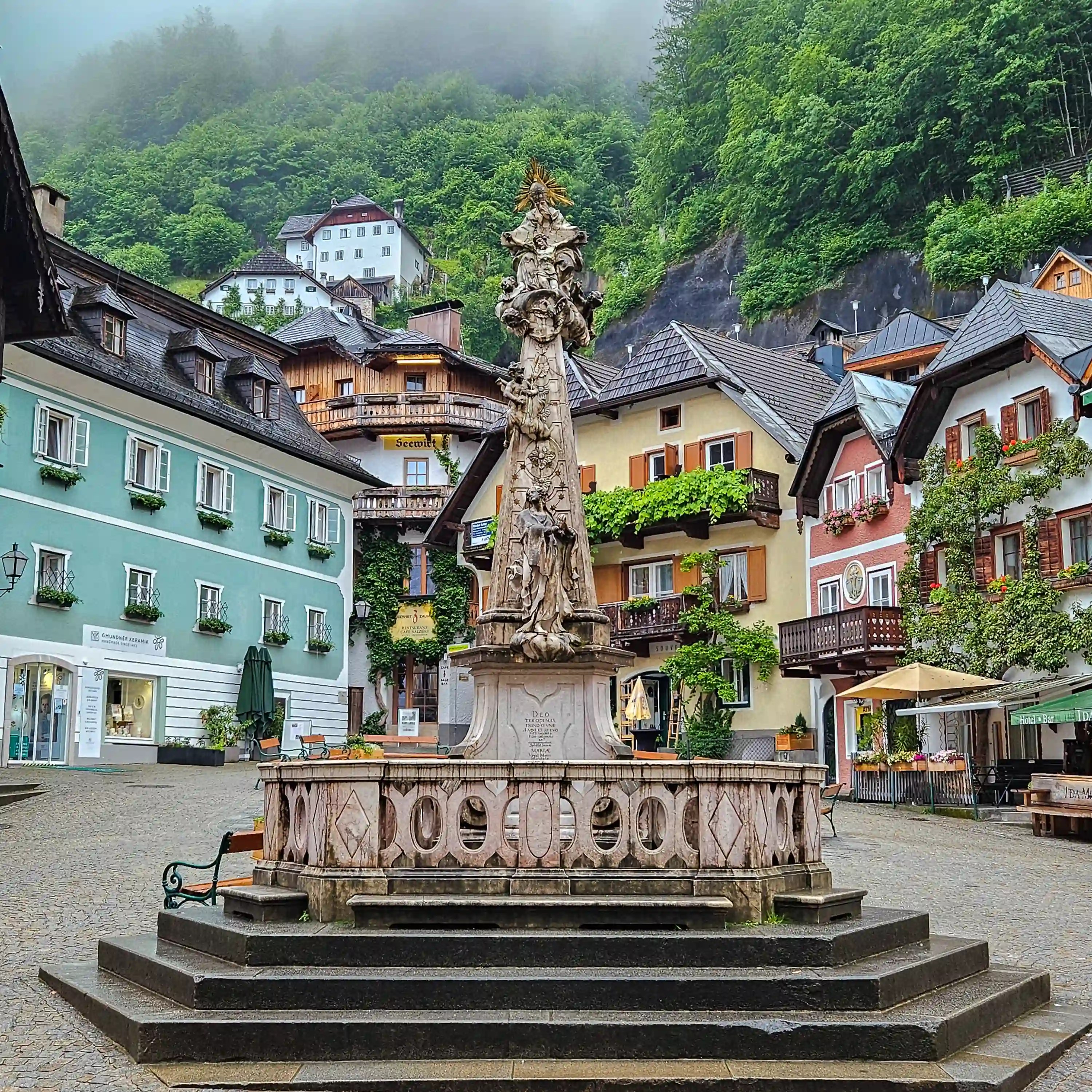 The Holy Trinity Column rises at the center of Hallstatt’s square with traditional houses and balconies behind it.