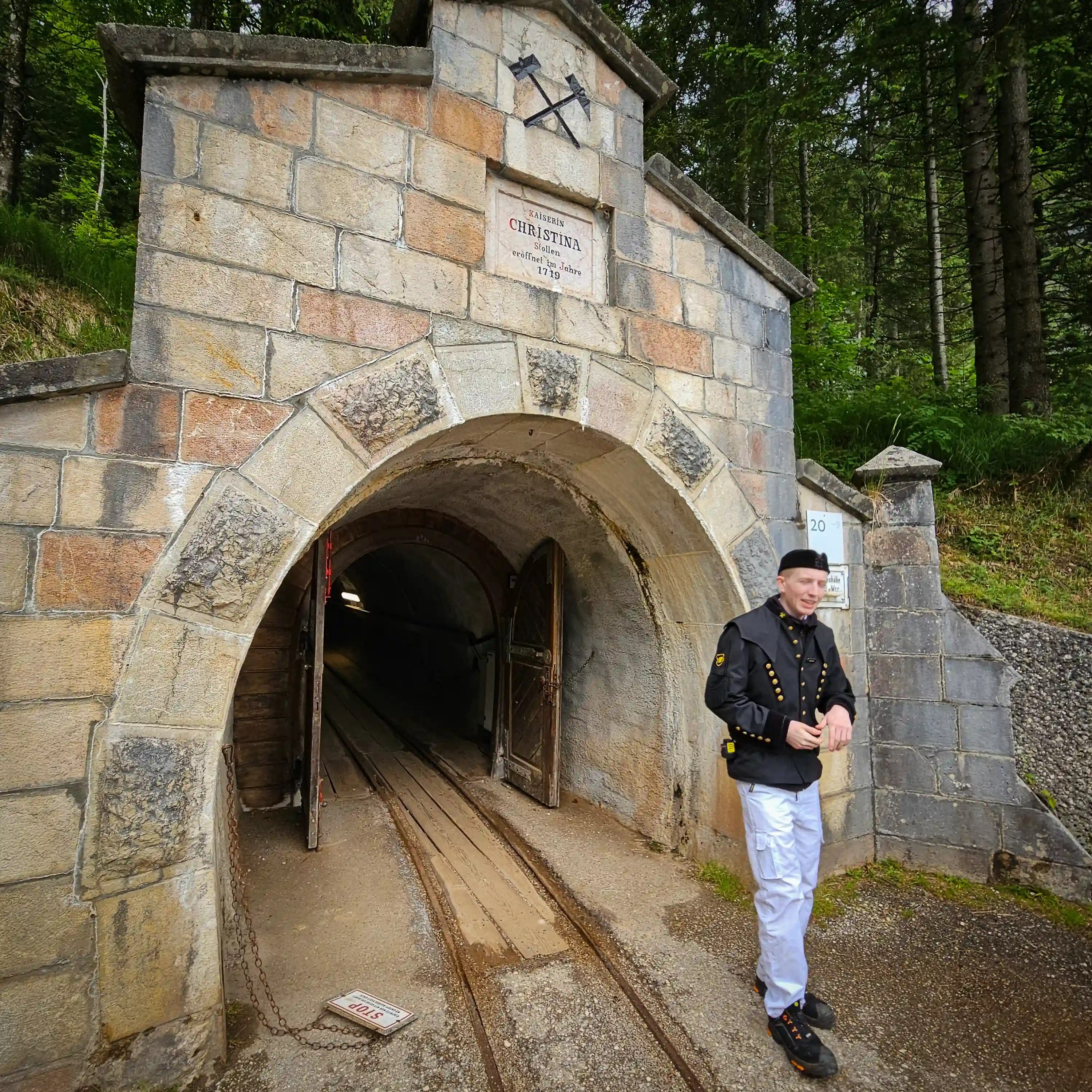 Stone mine portal at Hallstatt Salt Mine with open wooden doors, narrow rail tracks leading inside, and a guide in a miner’s jacket standing at the entrance.