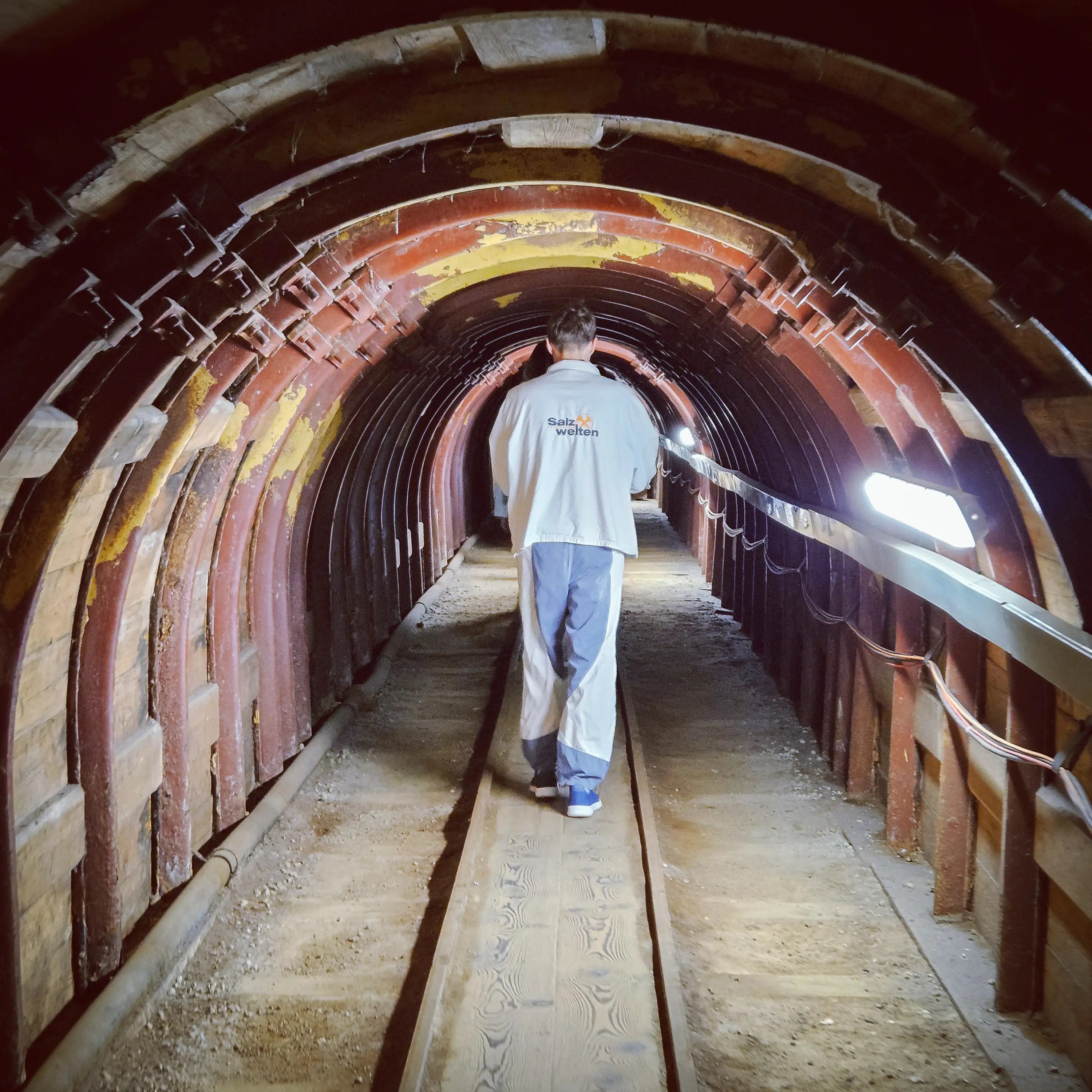 A dimly lit mining tunnel reinforced with arched metal ribs and a wood-plank central path, with a person walking away from the camera toward deeper darkness.