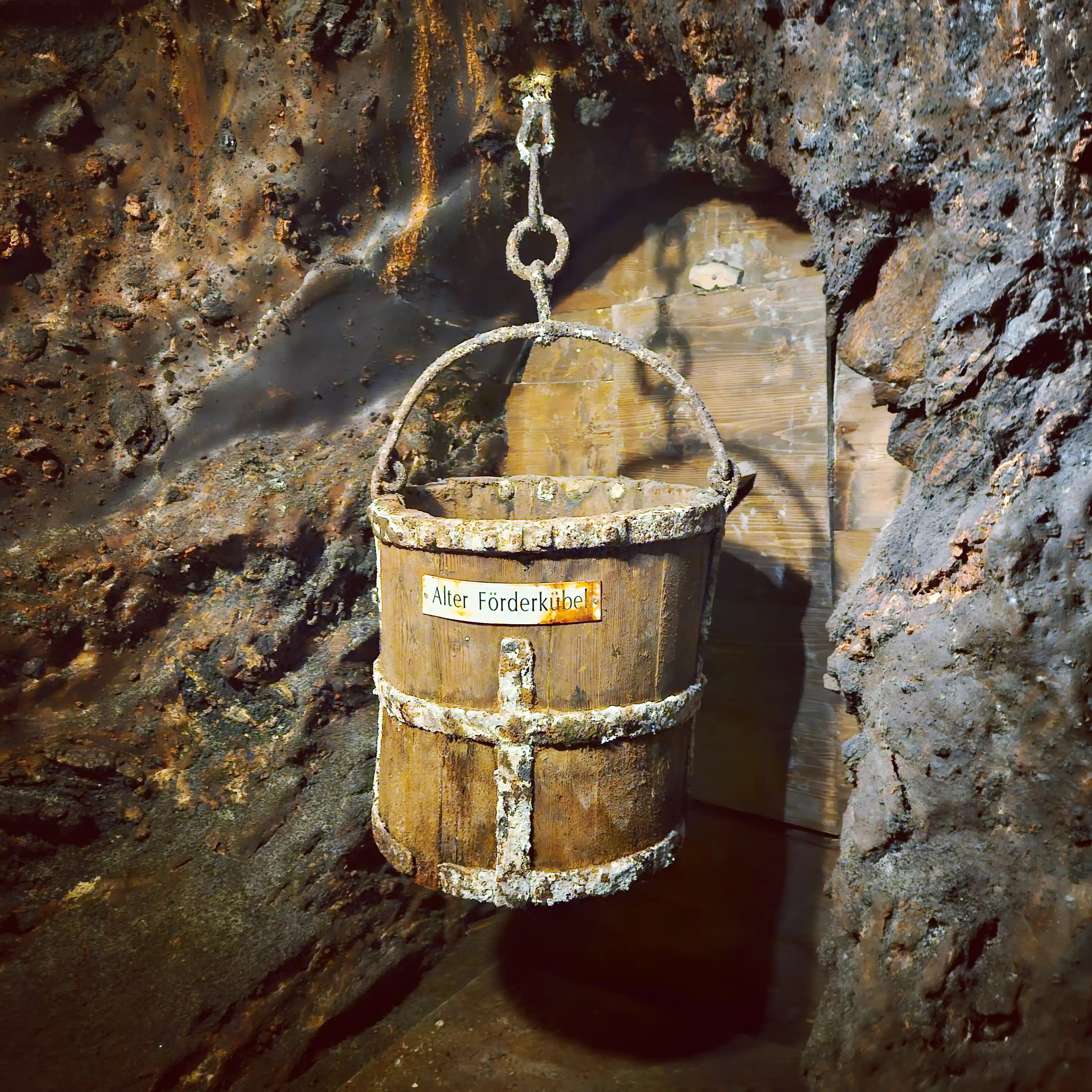 A wooden bucket labeled “Alter Förderkübel” suspended by a metal hook against a rough rock wall inside the salt mine.