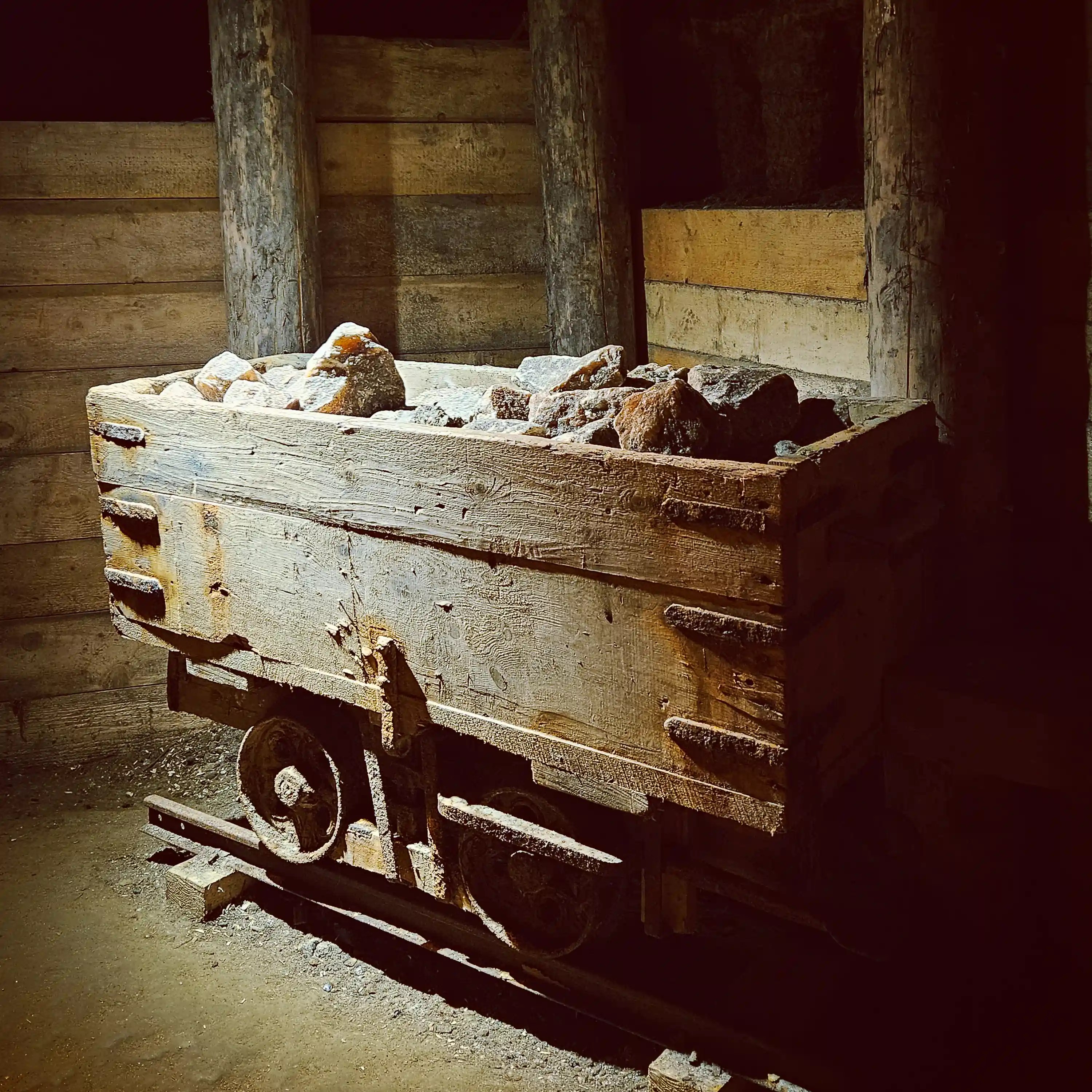 A weathered mine cart on narrow rails filled with rocks, positioned beside wooden walls inside the salt mine.