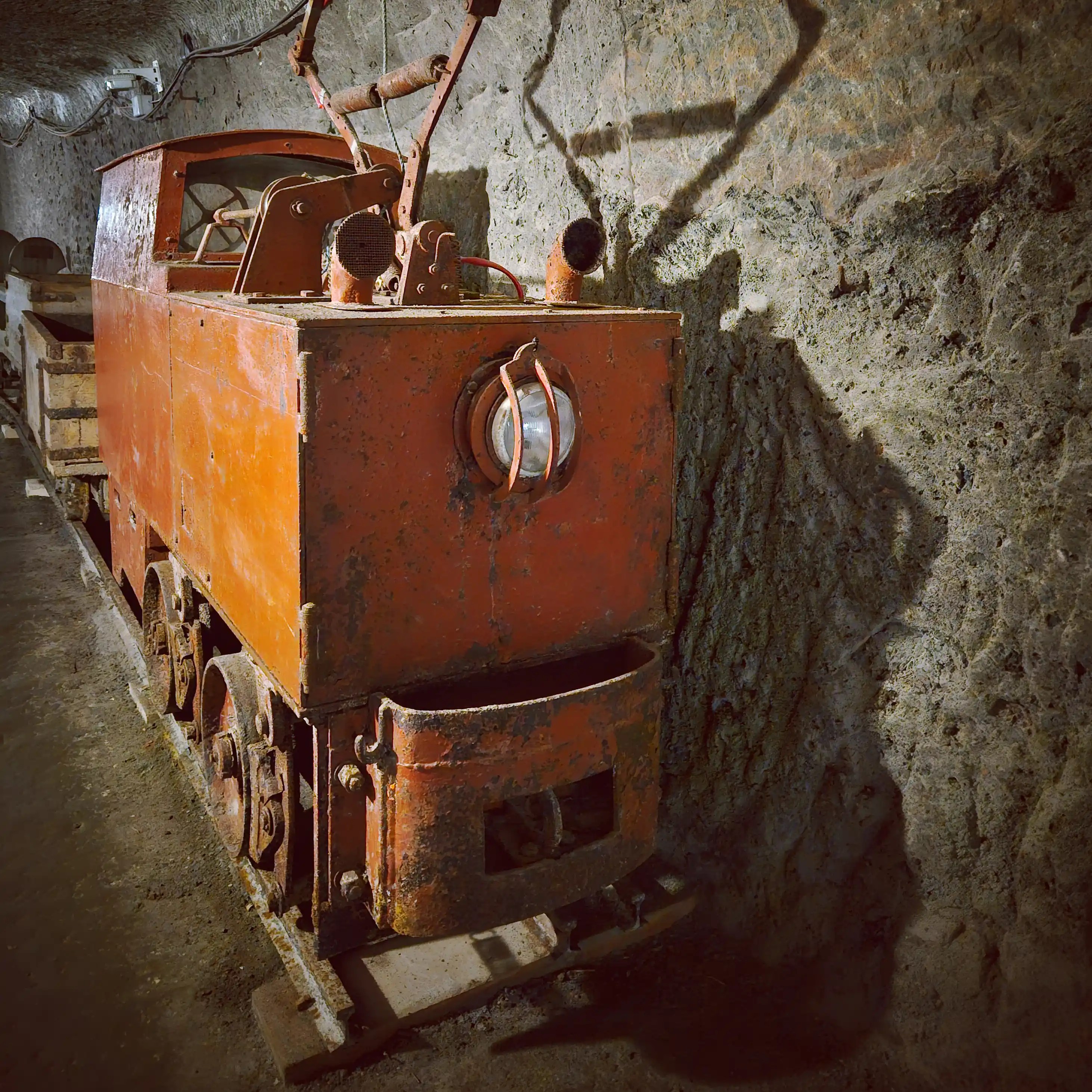 A small industrial locomotive with a headlamp and carts behind it parked on rails inside the salt mine.