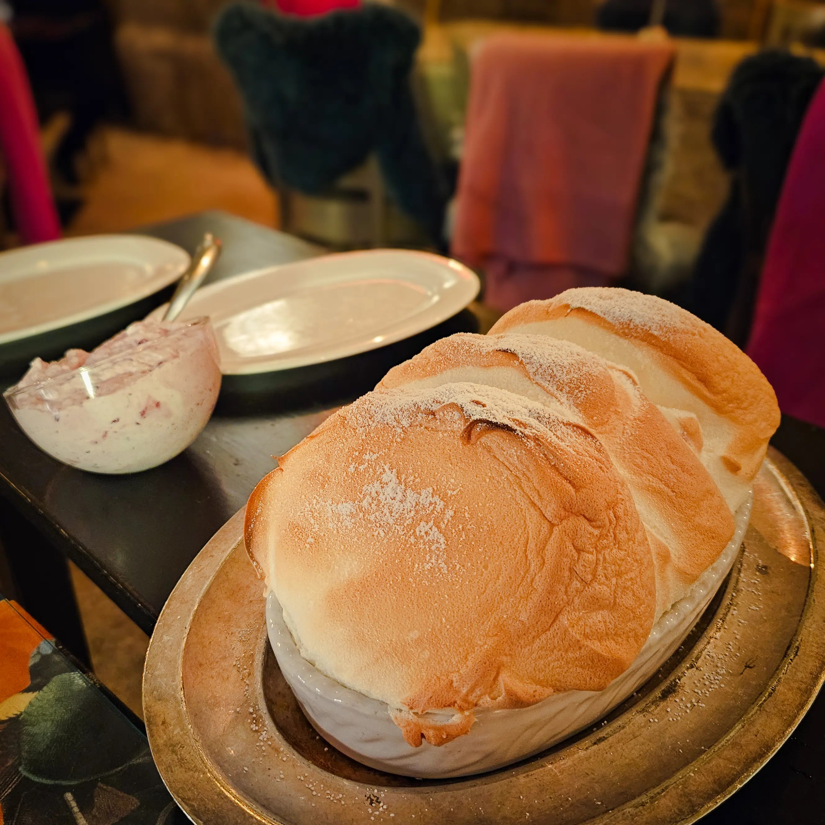 A large Salzburger Nockerl dessert dusted with powdered sugar sits in a white dish on a metal platter at a restaurant table.
