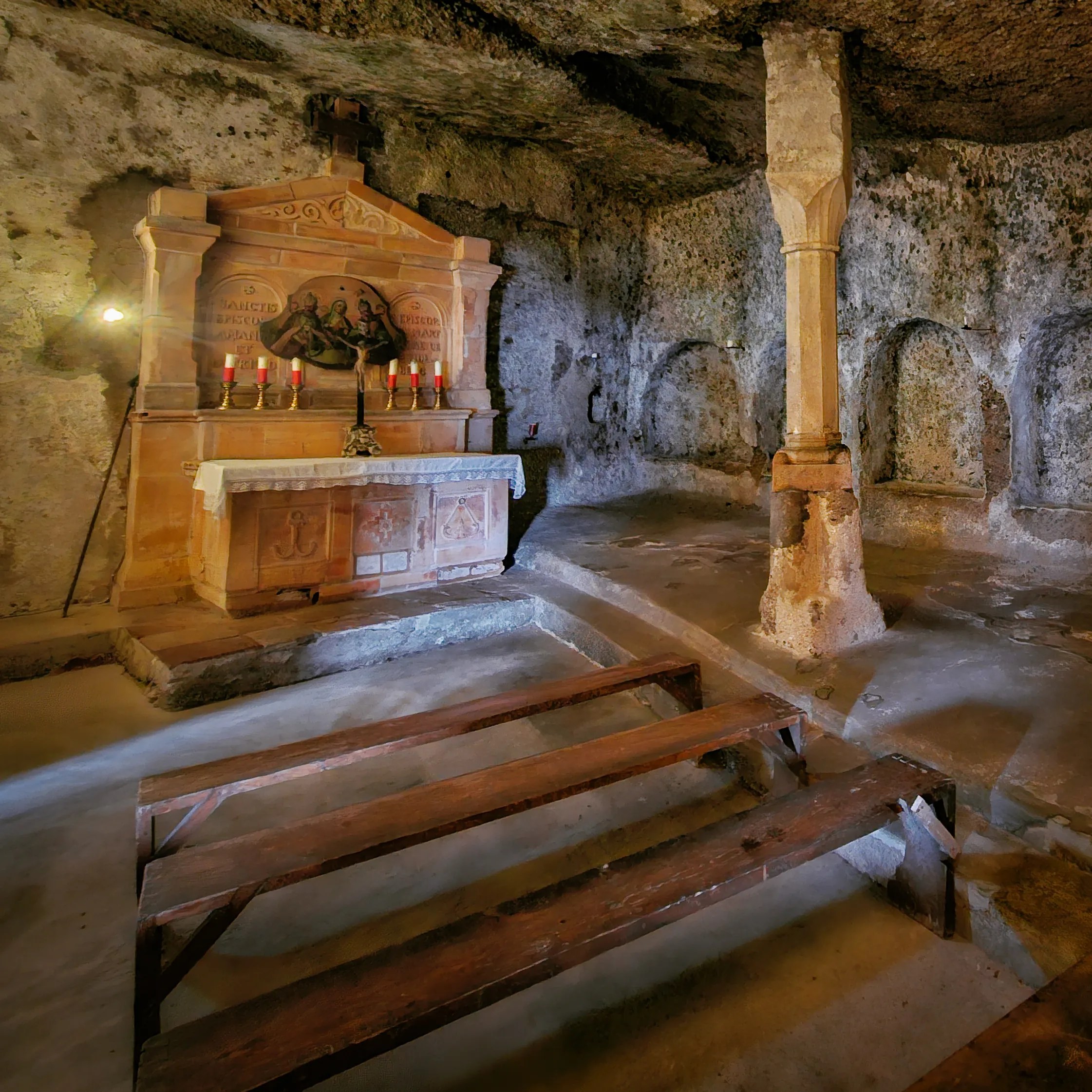 A carved stone altar with candles and wooden benches sits inside a rock-cut chamber in the St. Peter’s Catacombs.