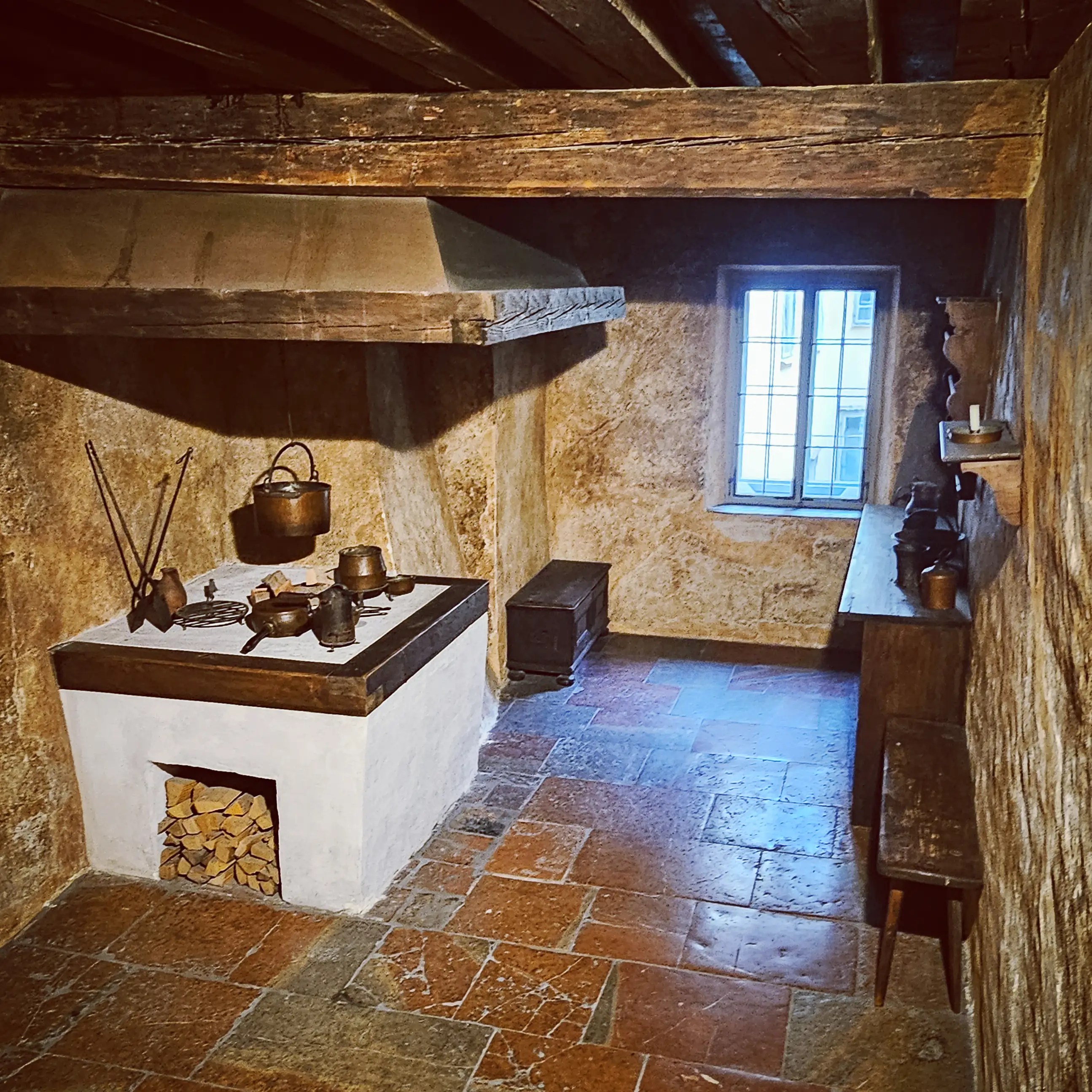 A historic kitchen inside Mozarts Geburtshaus with a white masonry stove, hanging copper cookware, wooden beams, and a small window letting in daylight.