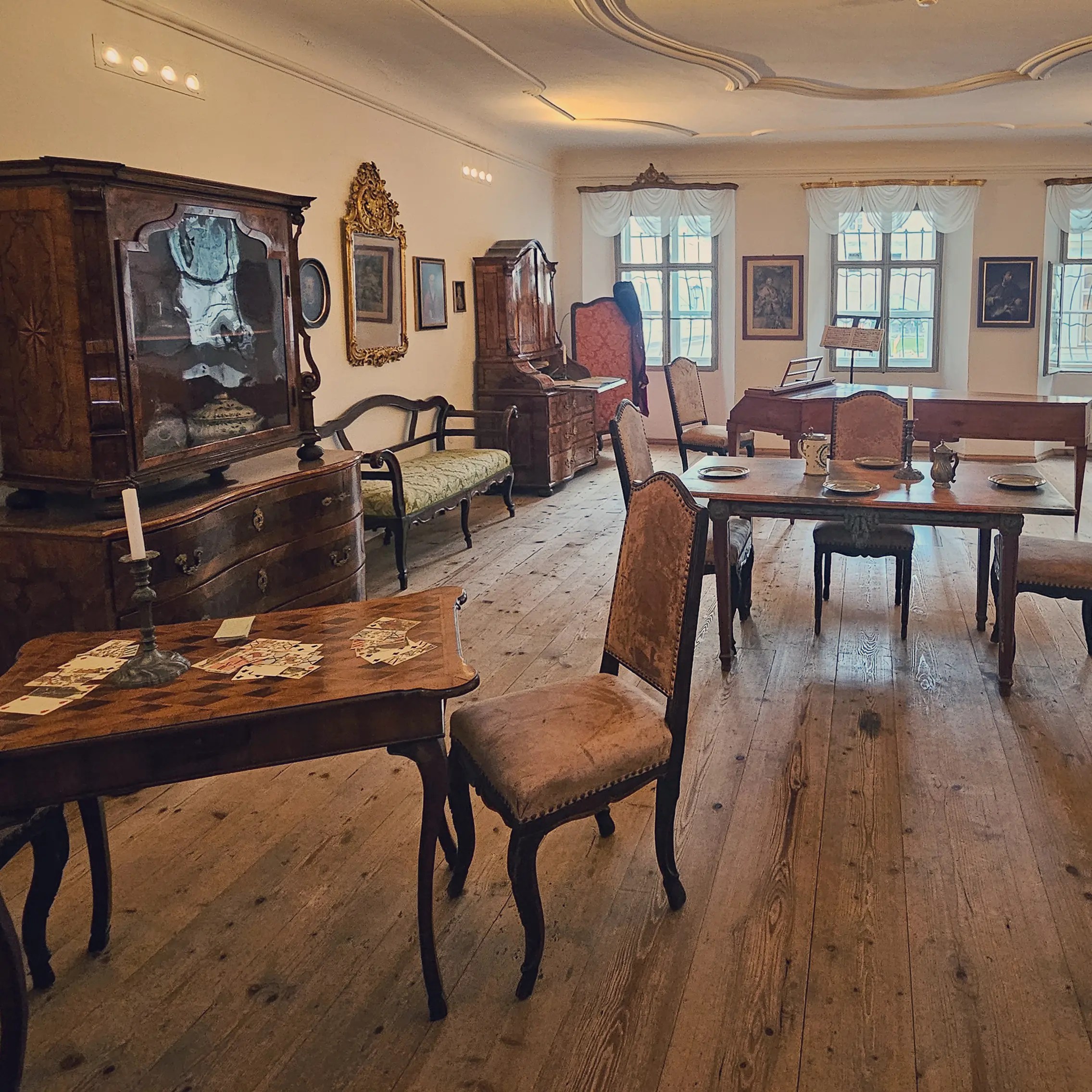 A furnished historic room inside Mozarts Geburtshaus with wooden floors, antique chairs, tables set with dishes, framed artwork, and large windows along the far wall.