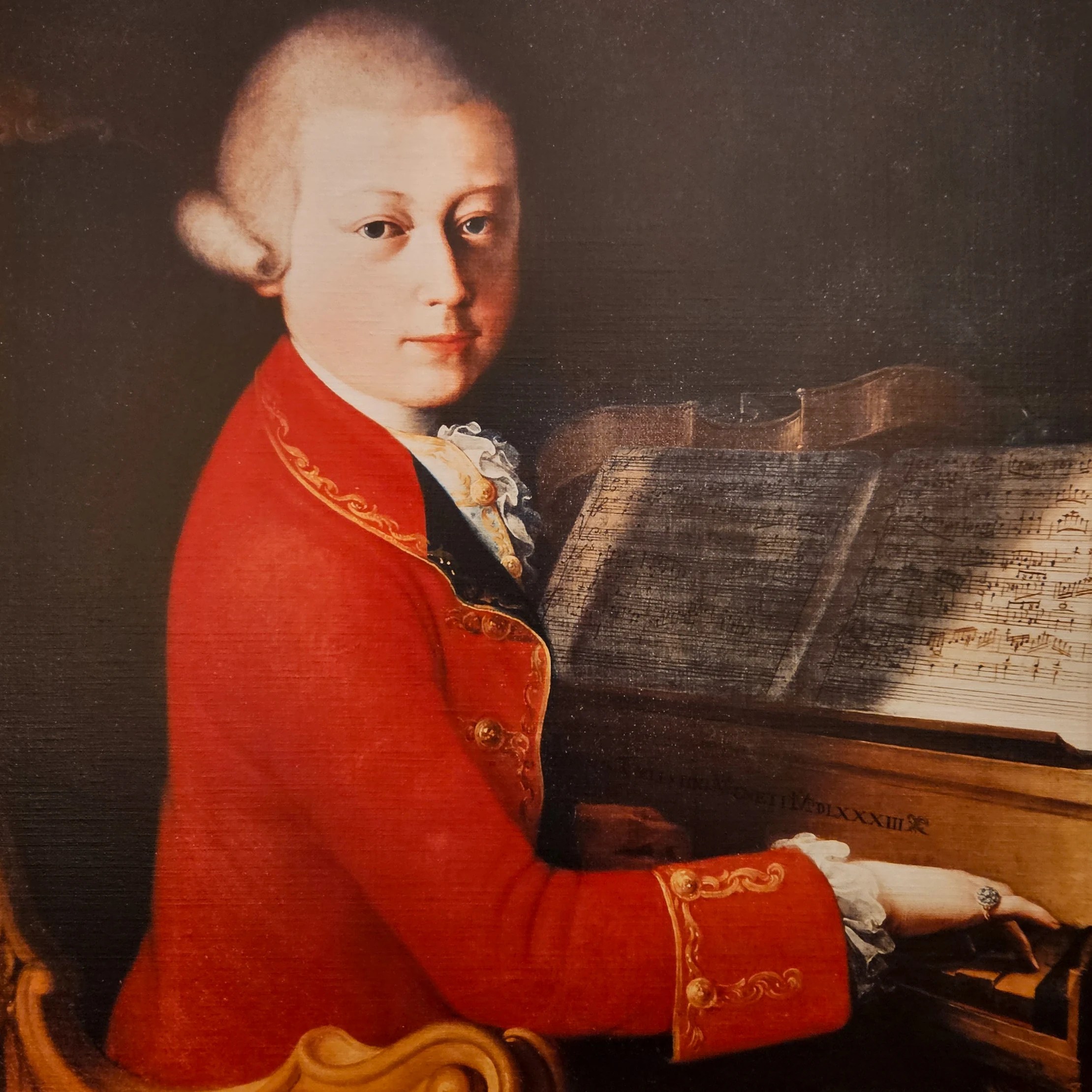A copy of a painted portrait of a young Wolfgang Amadeus Mozart wearing a red coat while seated at a keyboard with sheet music in front of him.