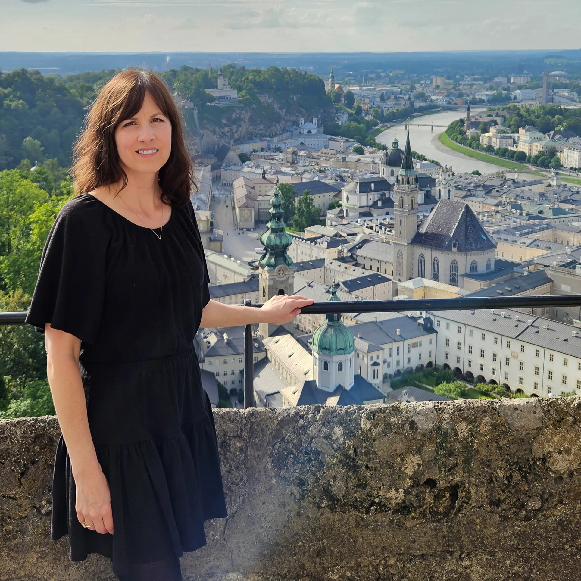A woman in a black dress stands at a stone railing overlooking Salzburg’s Old Town, with church domes, towers, and the Salzach River below.
