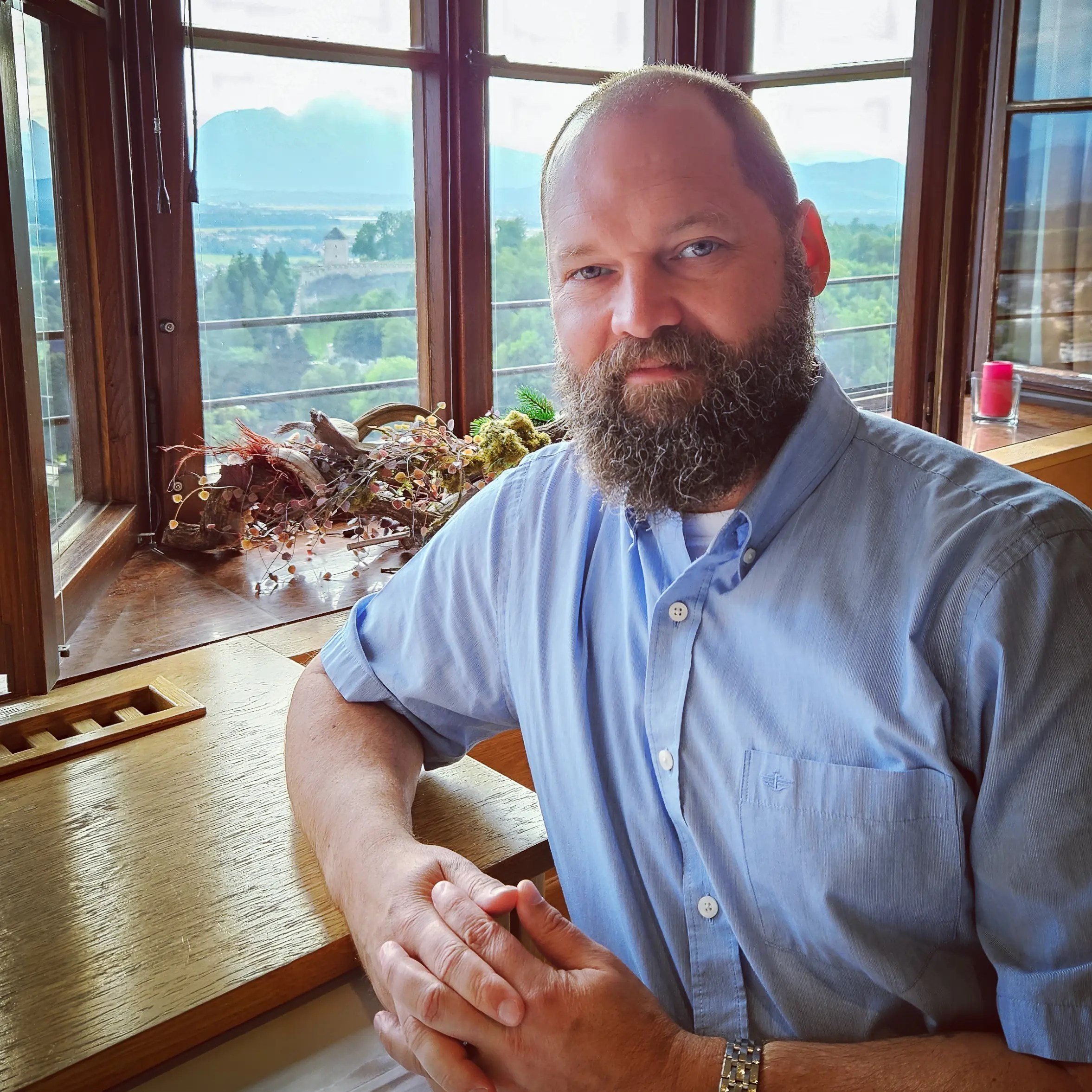 A bearded man seated at a wooden table inside the fortress dining room, with windows and a mountain landscape behind him.