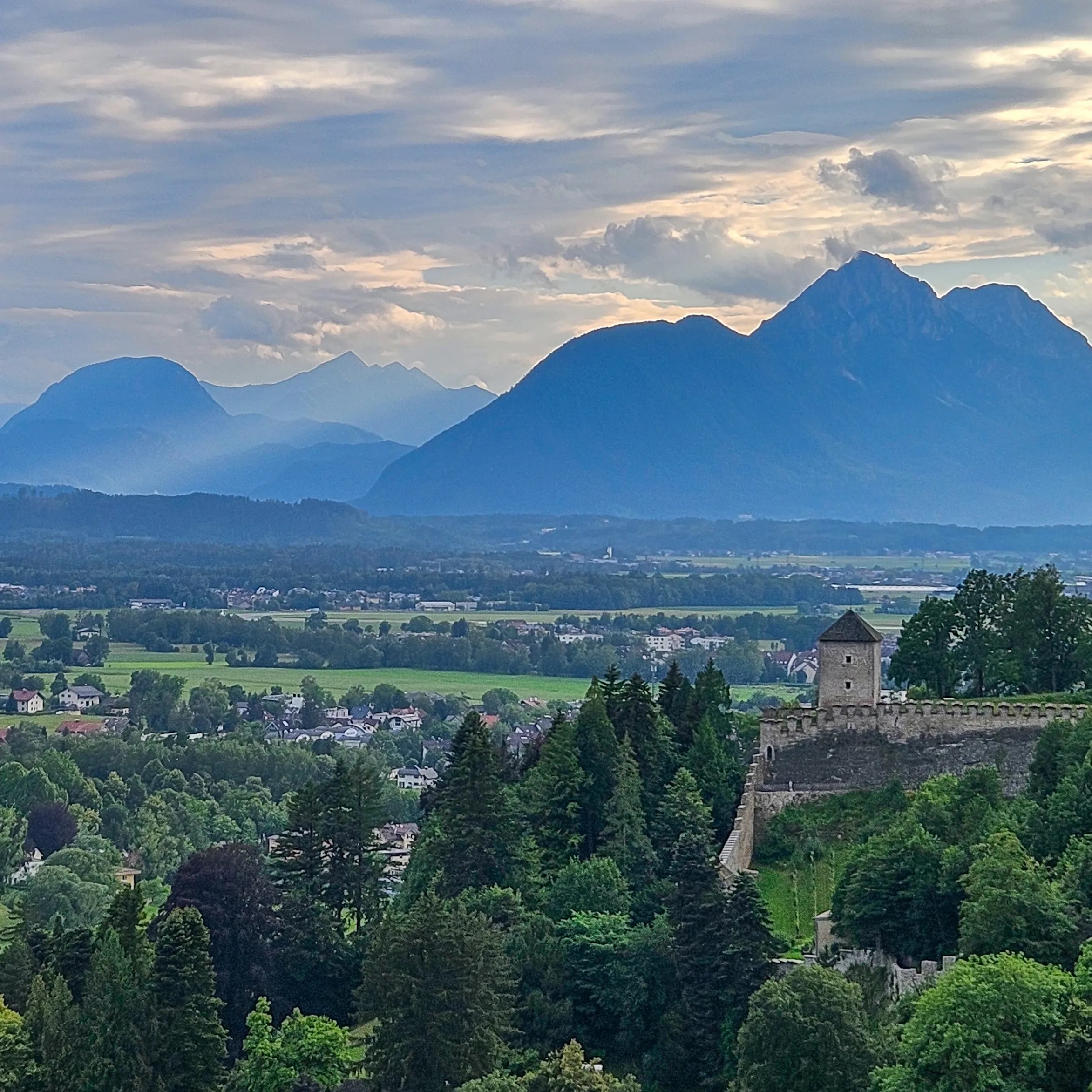 A stone watchtower and fortress wall rise above dense green trees, with layered blue mountains in the distance.