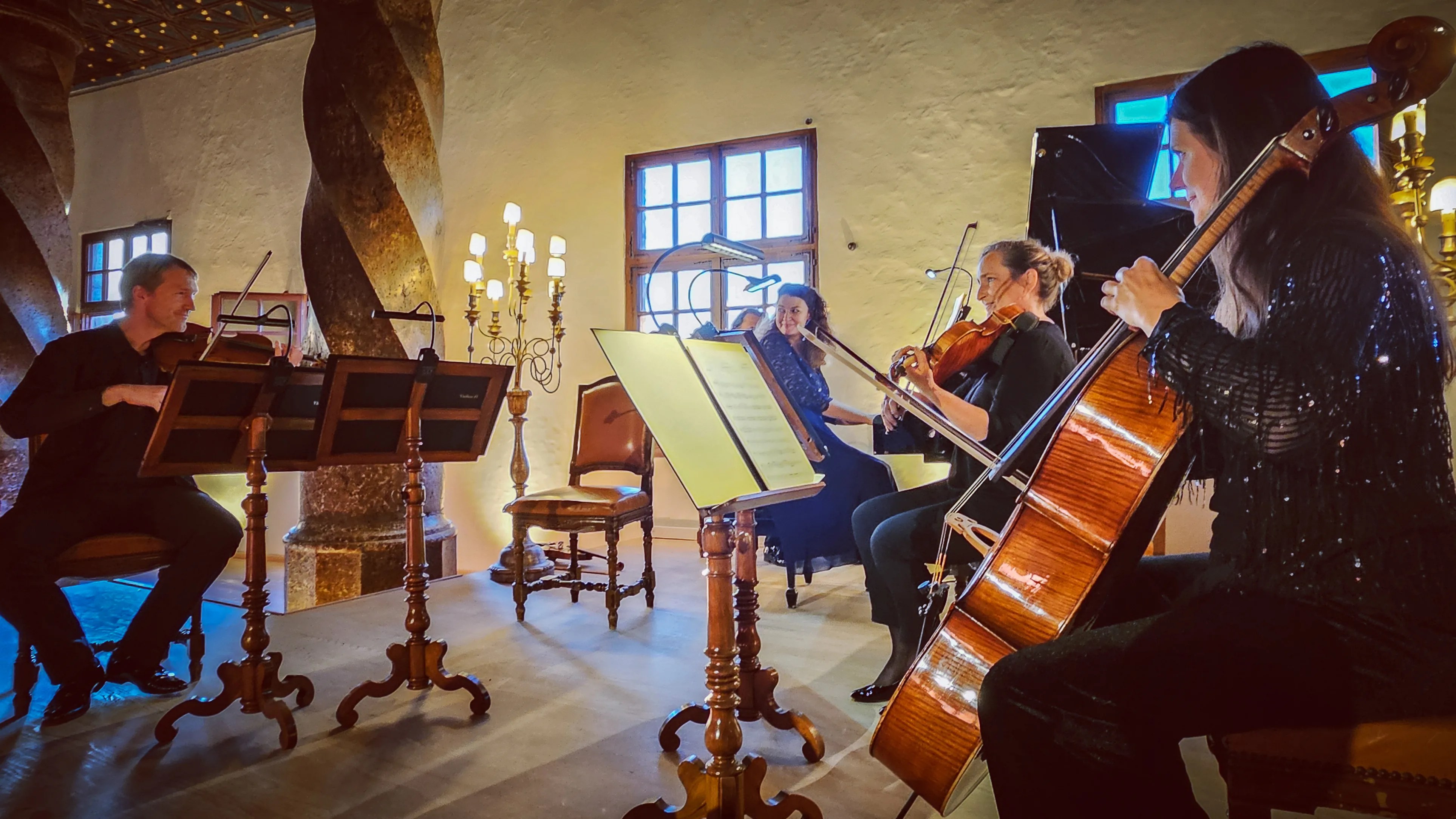A small classical ensemble performs in an ornate hall, with musicians playing violin, cello, and piano beneath a decorated ceiling.