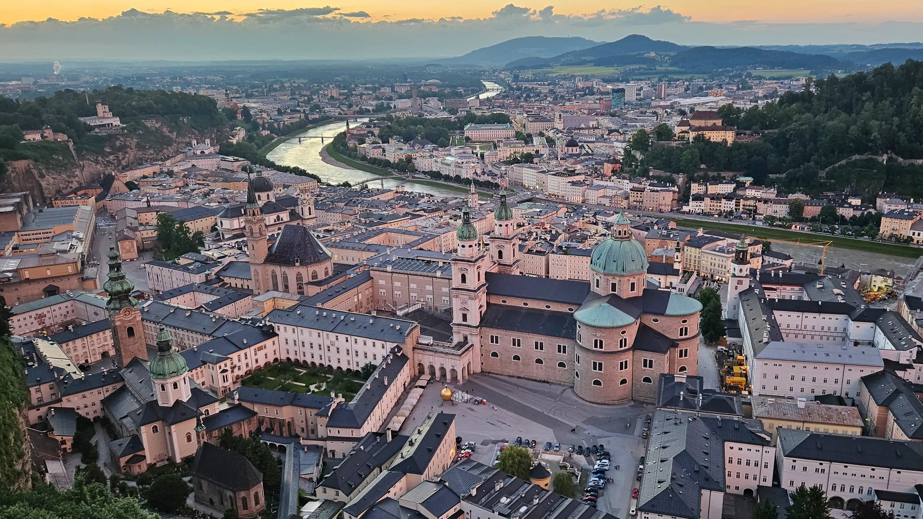 An aerial view of Salzburg at sunset, with the Salzburg Cathedral and surrounding Old Town buildings along the Salzach River.