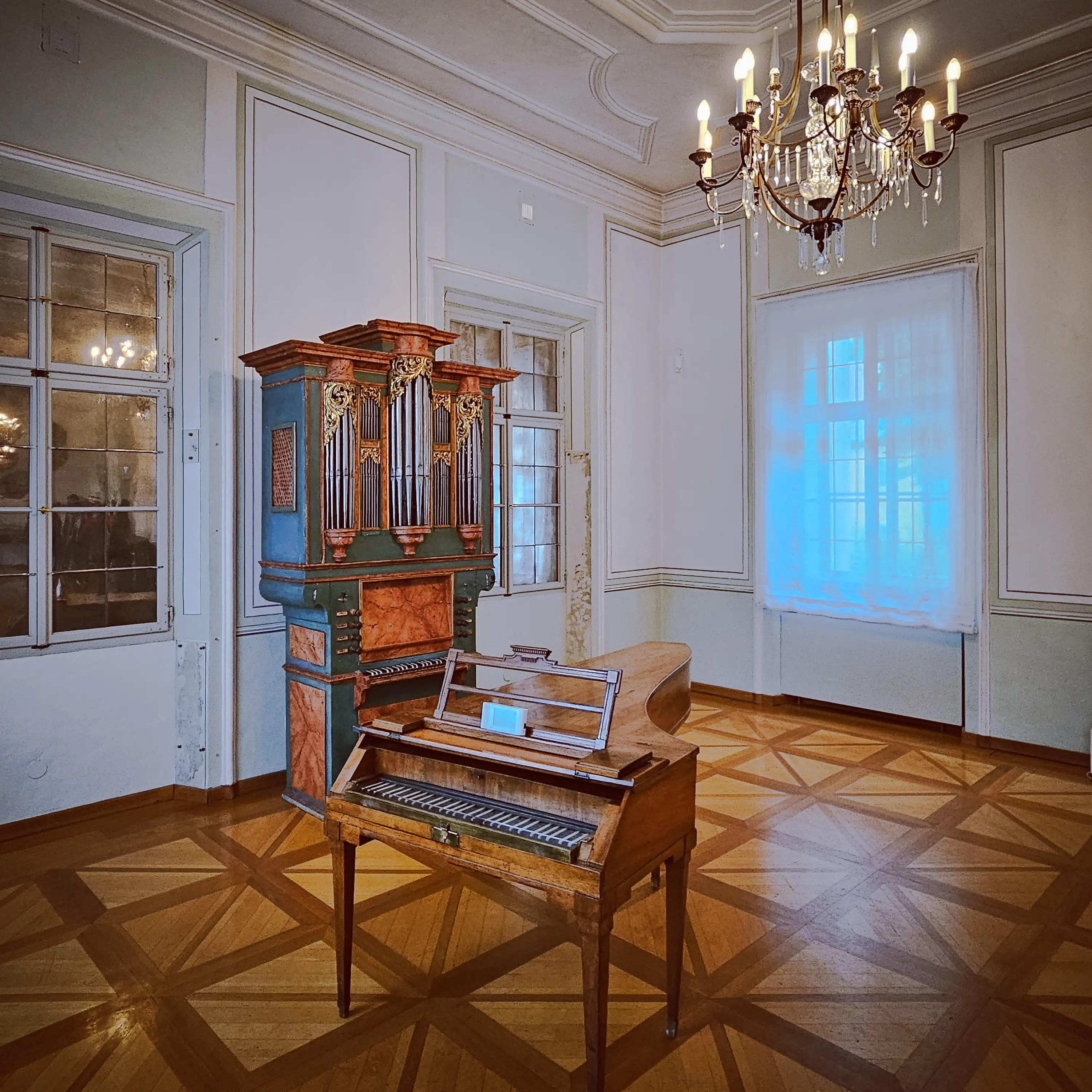 A historic music room inside Mozarts Geburtshaus featuring a small keyboard instrument, an ornate pipe organ, parquet flooring, and a chandelier overhead.