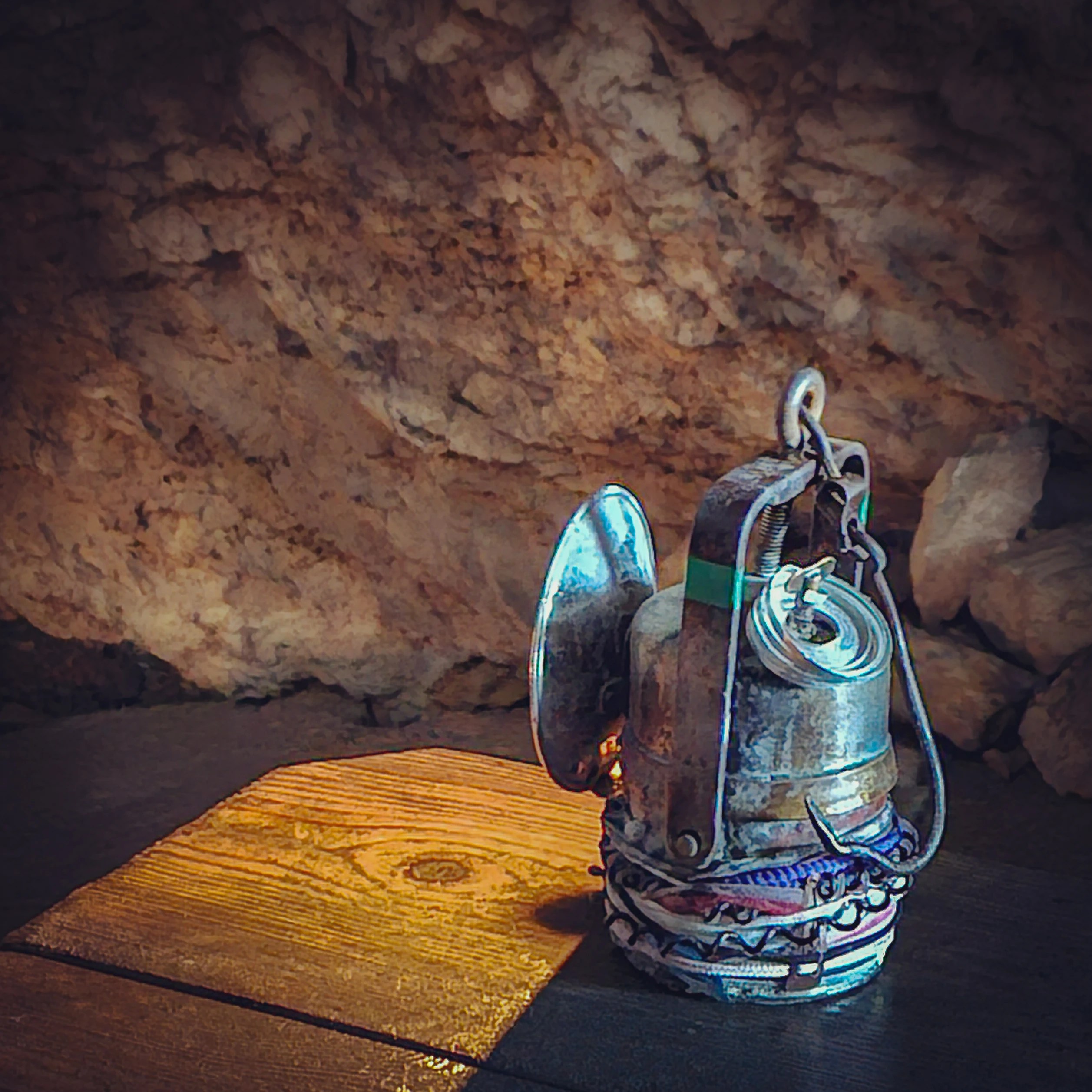 A metal miner’s lantern resting on a wooden surface inside a cave, casting warm light against rough stone walls.