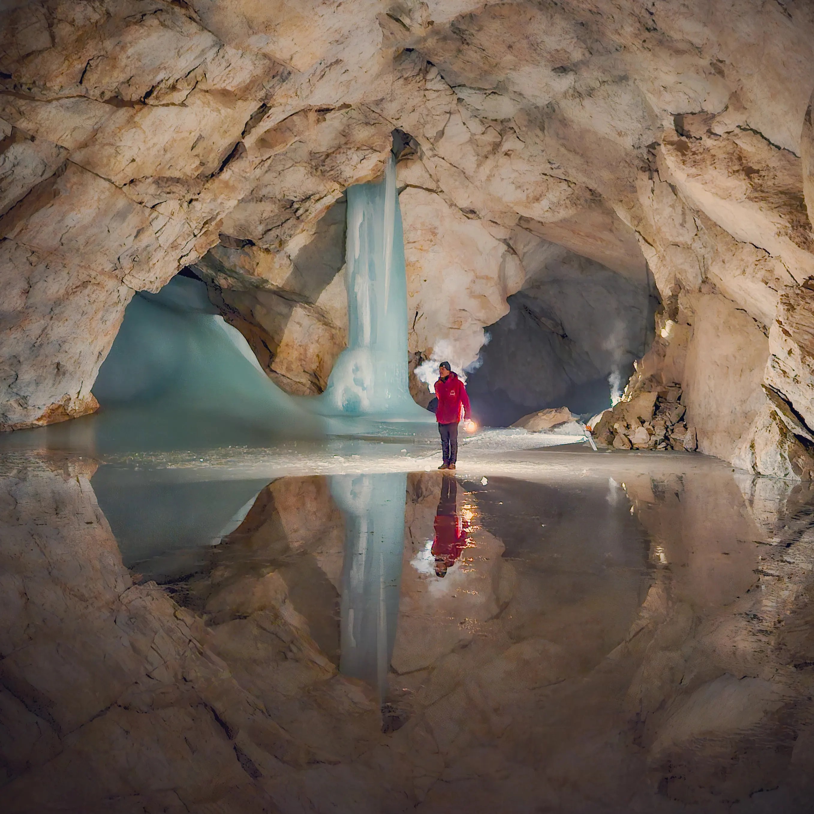 A guide holding a flare stands in a large ice chamber at Eisriesenwelt, reflected in shallow water beneath towering ice columns.