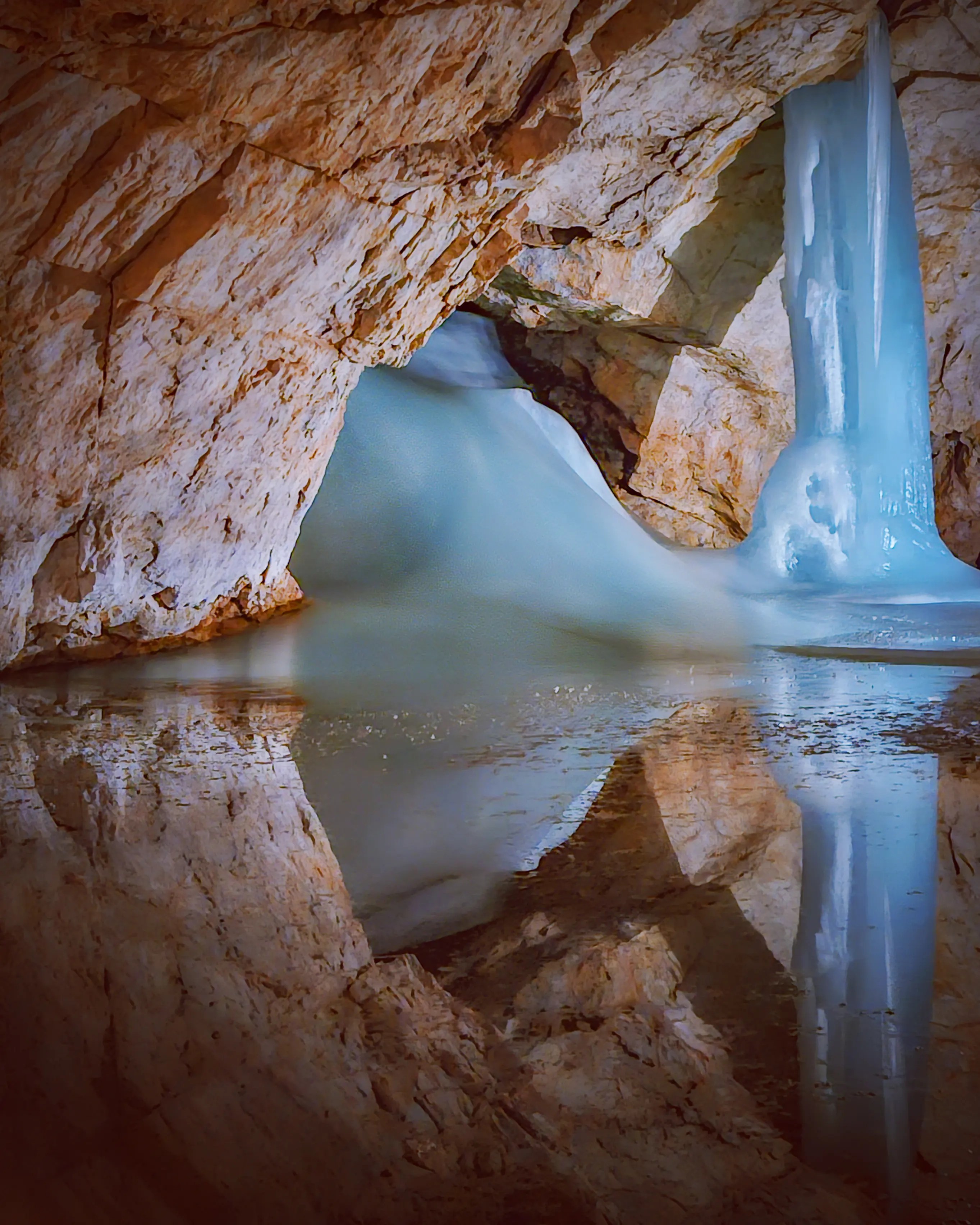 A smooth blue ice column descends from the cave ceiling into shallow water, reflecting against the rocky walls of Eisriesenwelt.