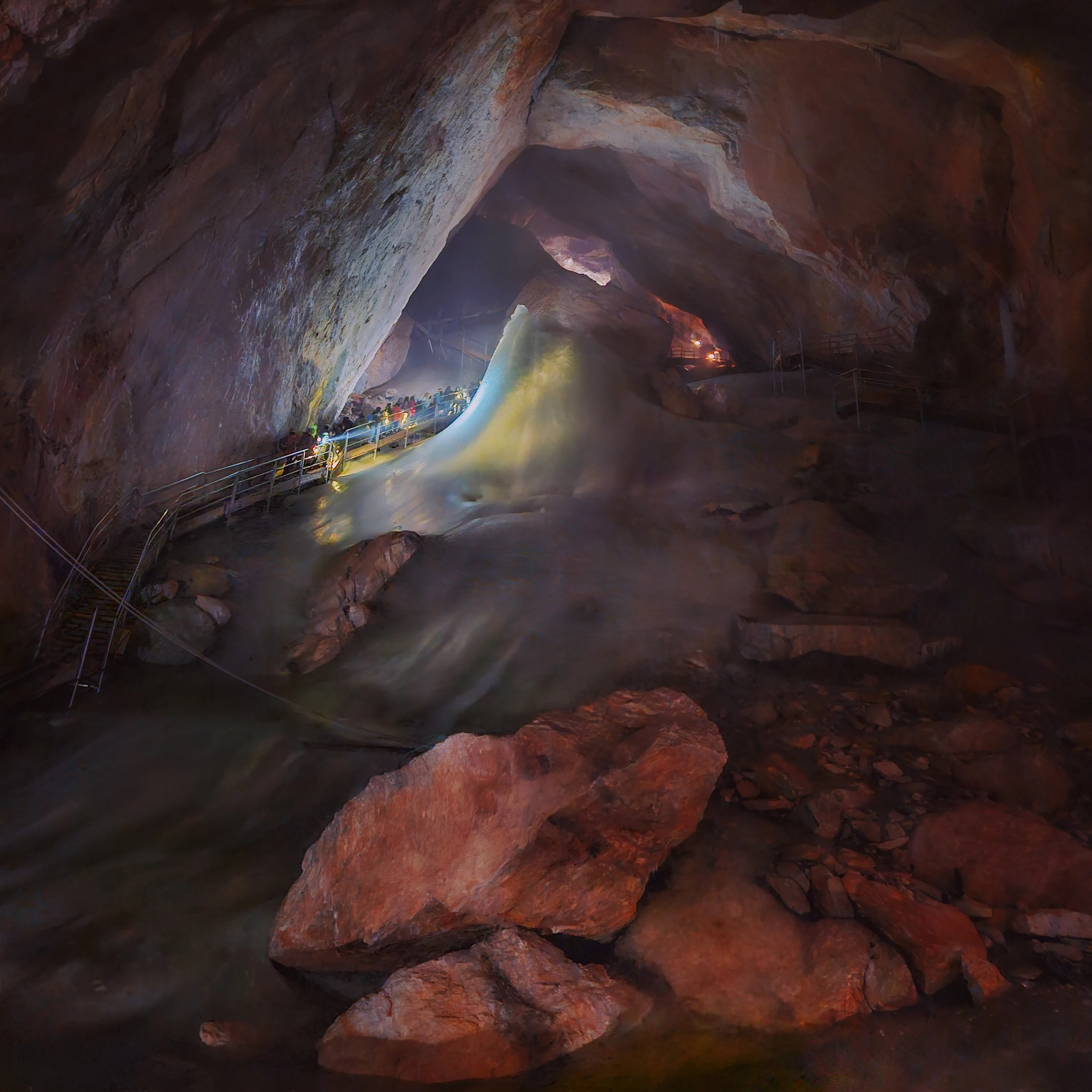 A wide view of an ice-filled chamber at Eisriesenwelt, with a glowing ice mound and visitors standing along a metal walkway.