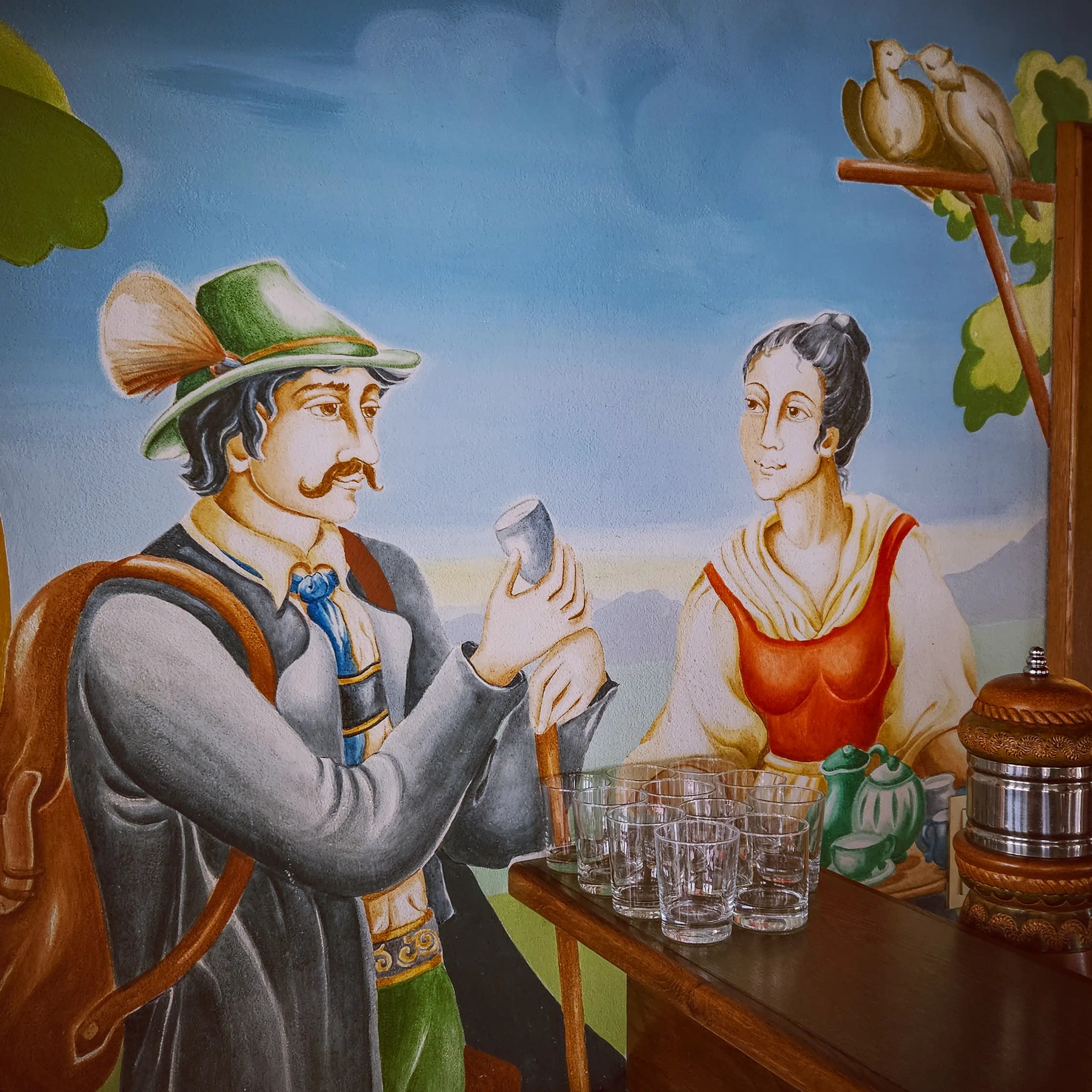 A painted mural showing a man in traditional Bavarian clothing holding a glass while a woman stands beside a table of glasses.