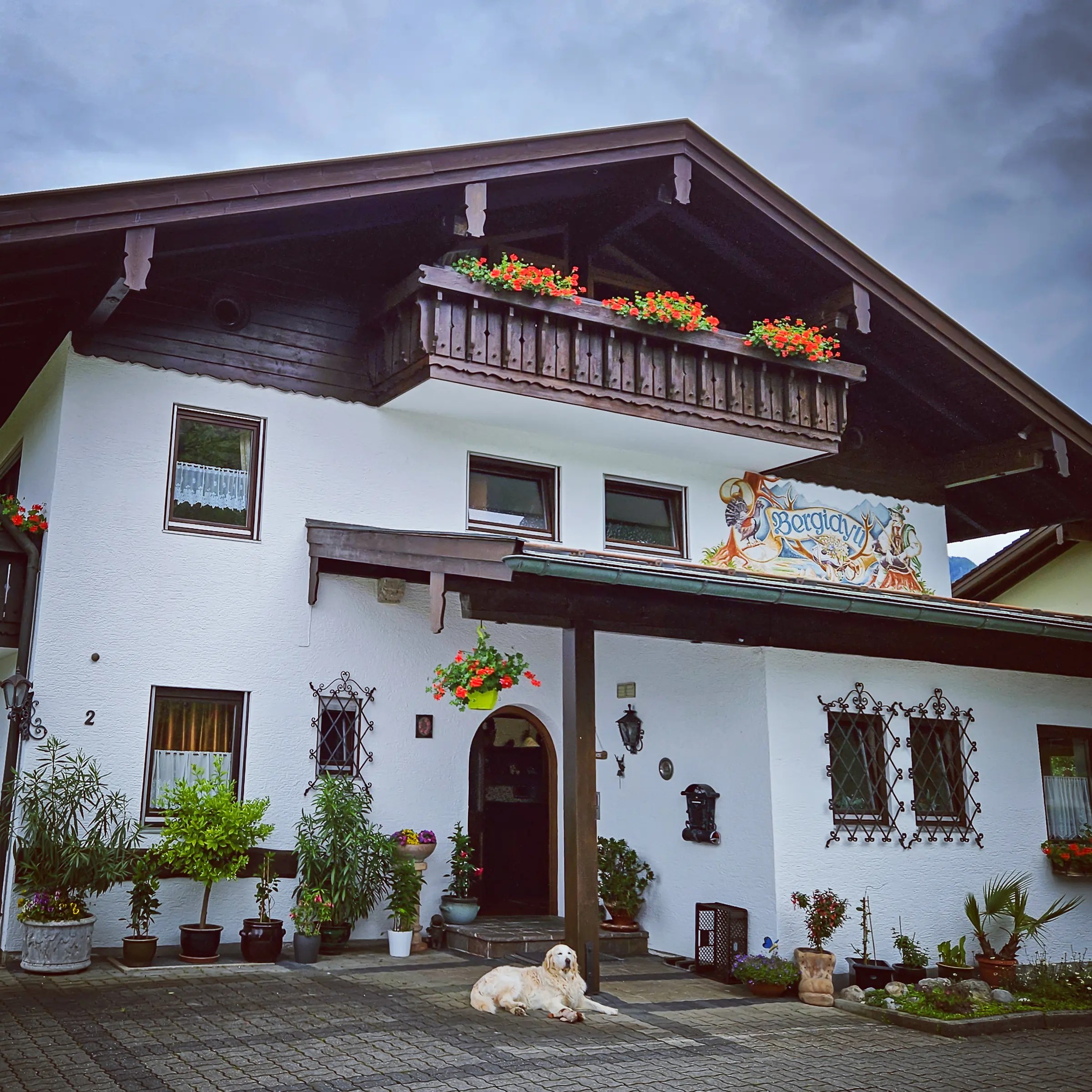 A white Alpine guesthouse with dark wooden balconies, flower boxes, and a painted sign above the entrance.