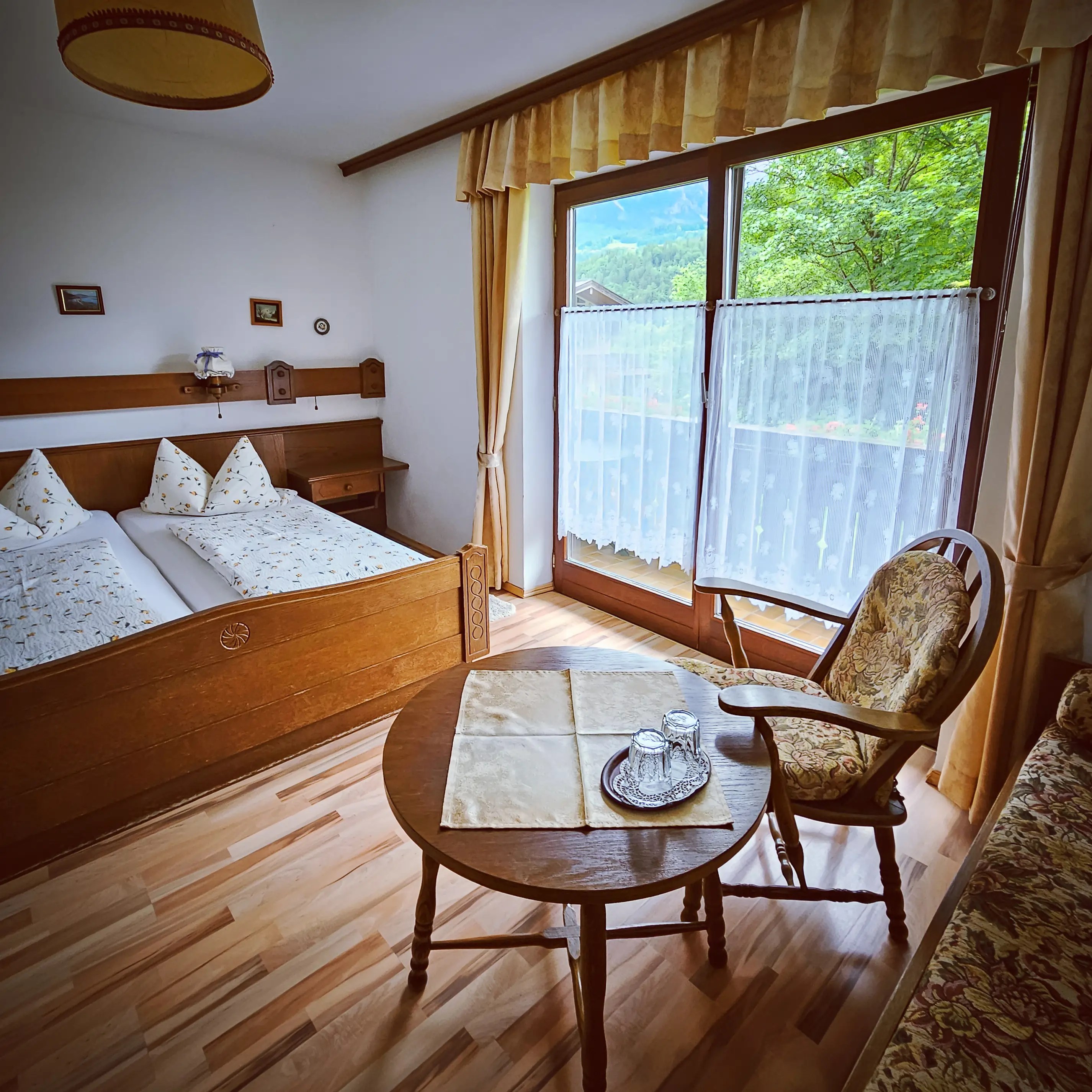 A bright guest room with a wooden bed, small sitting table, upholstered chair, and large windows overlooking greenery.