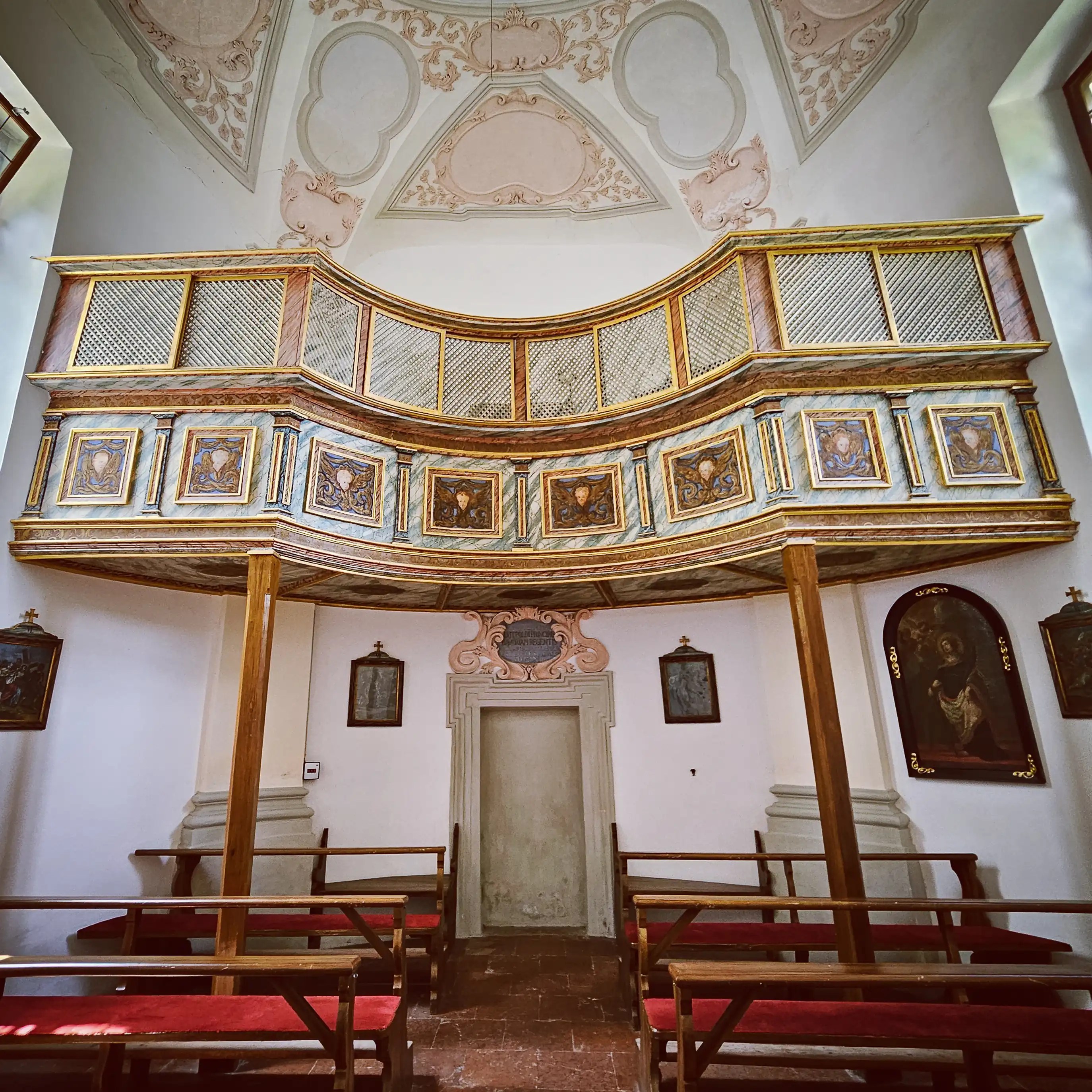 An ornate wooden gallery with painted panels curves above simple wooden pews inside the Church of St. Bartholomä.