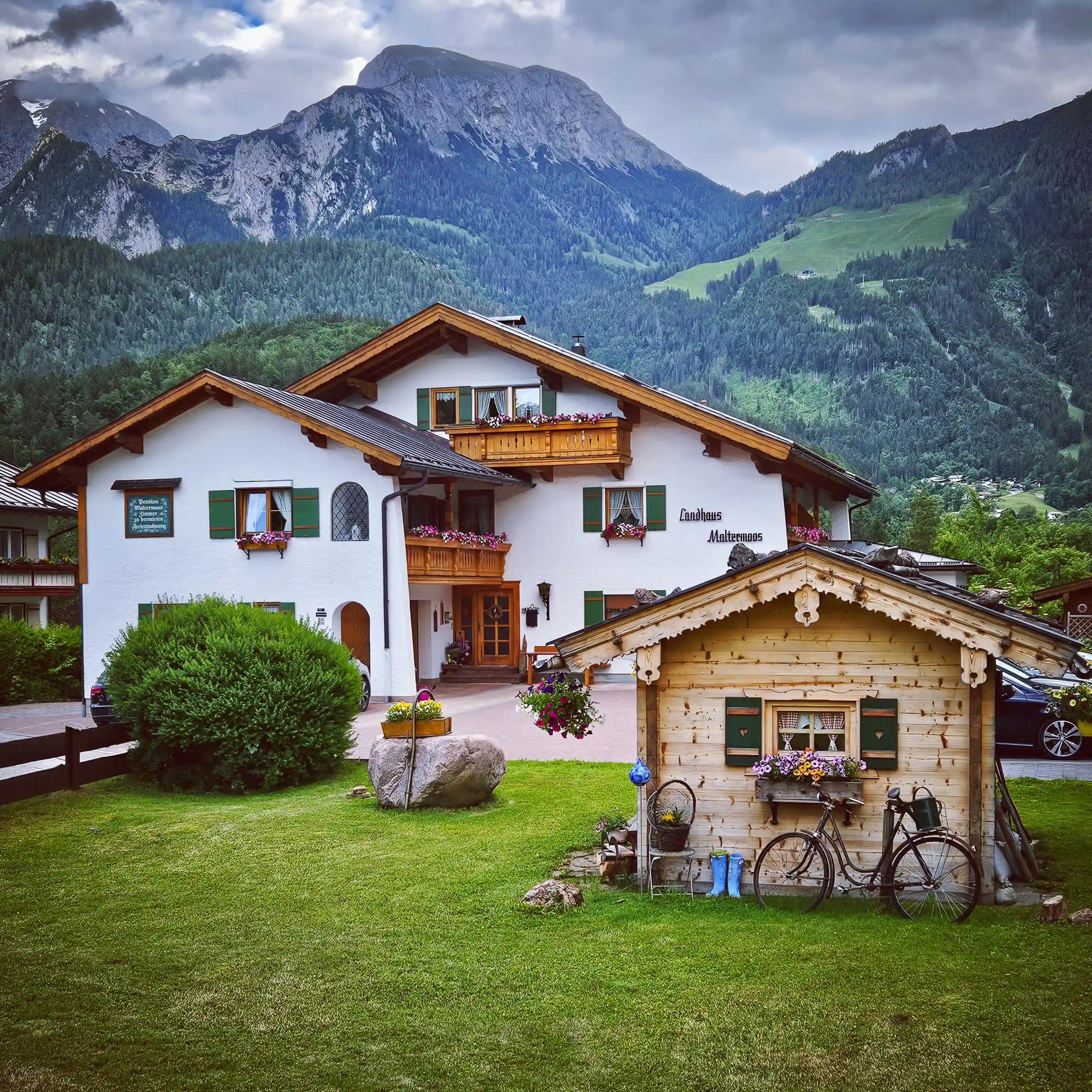 A white Alpine house with green shutters, flower-filled balconies, and a small wooden outbuilding set against mountain scenery.