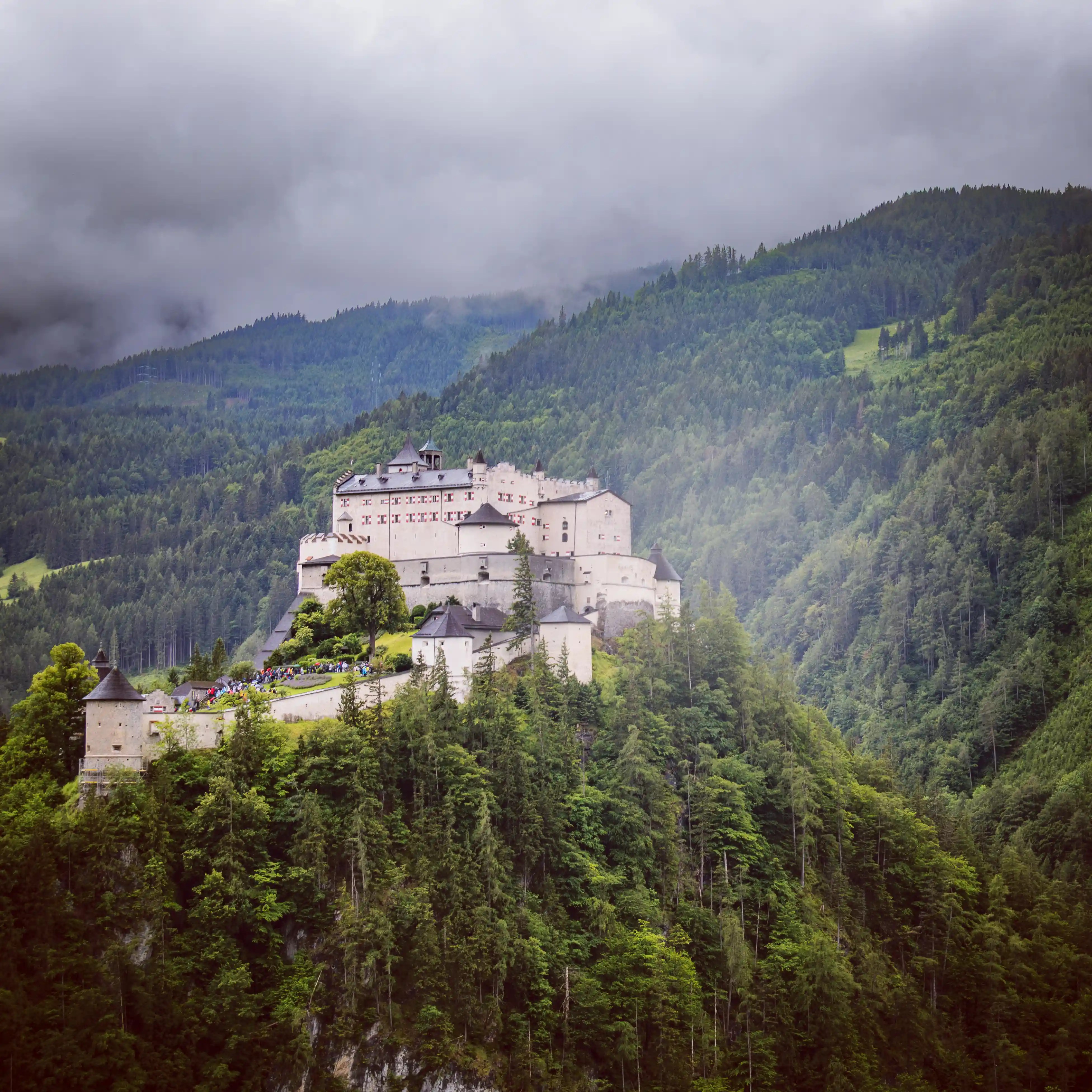 A view of Burg Hohenwerfen shows the fortress centered on a wooded hill with layered mountain slopes behind it.