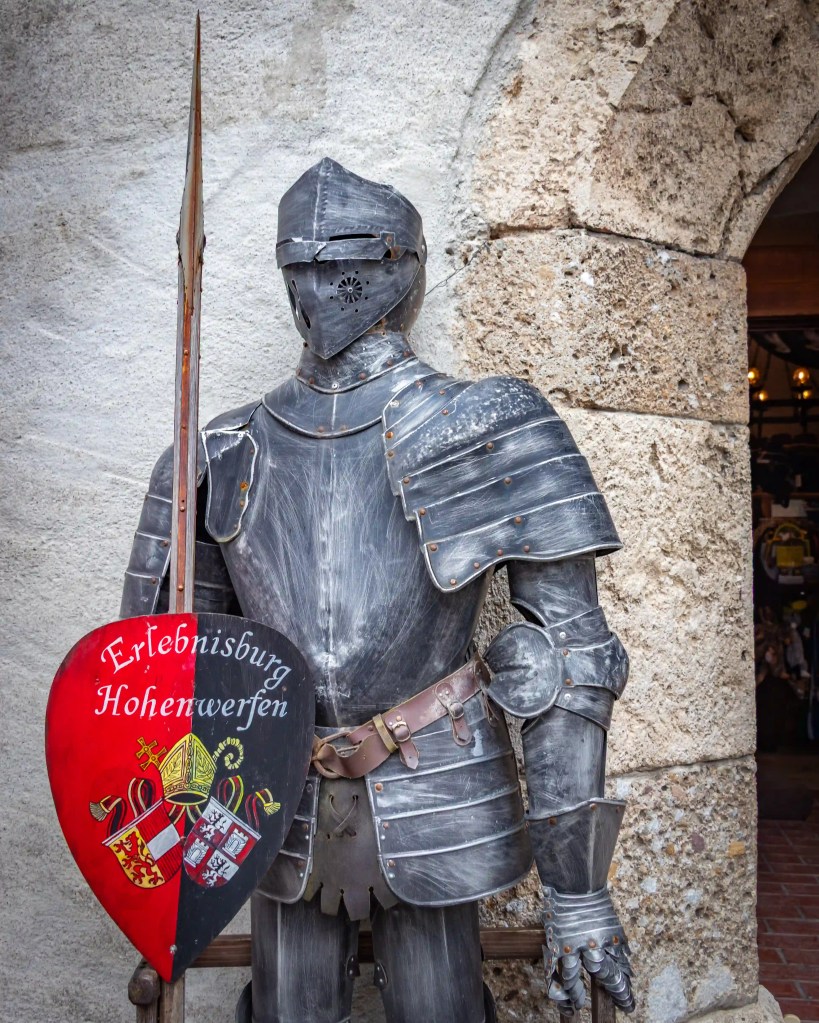 A full suit of medieval armor stands against a stone wall at Burg Hohenwerfen, holding a shield marked with the castle’s name.