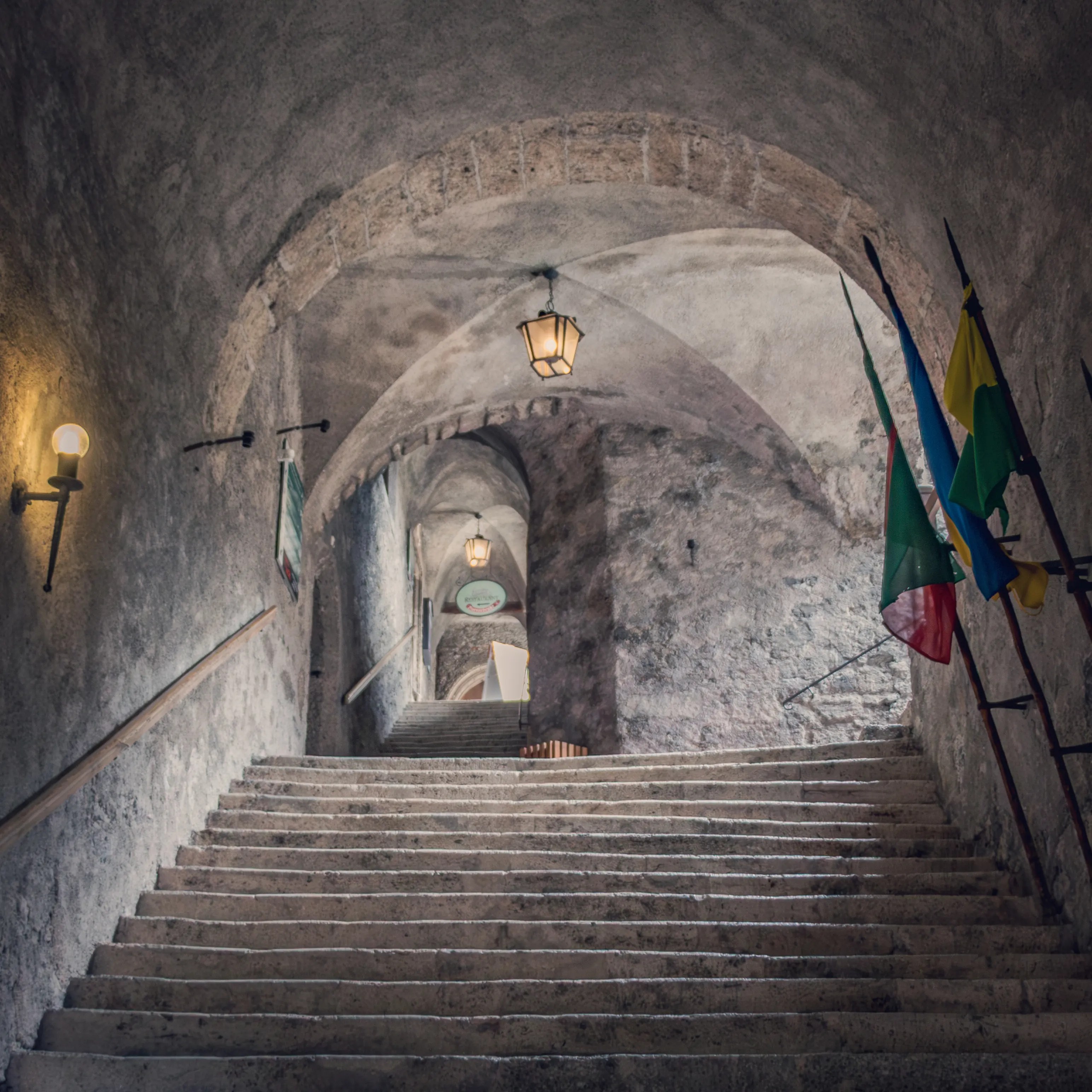 A long stone staircase curves upward through arched ceilings inside Burg Hohenwerfen, lit by hanging lanterns.