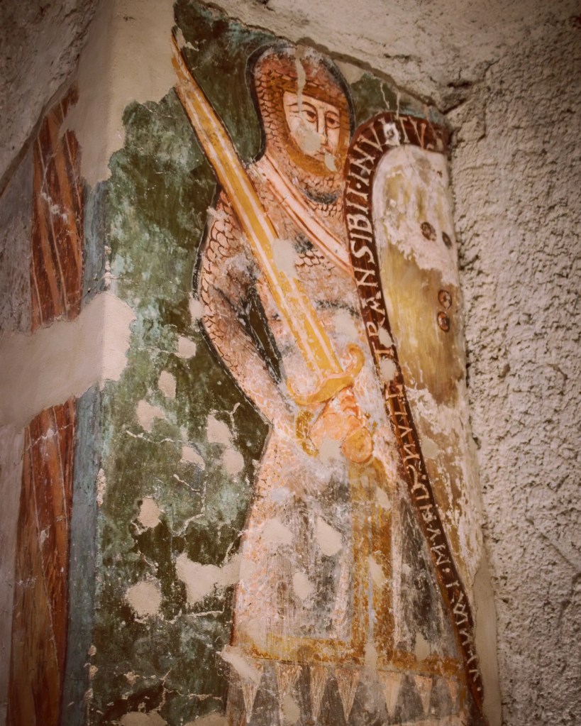 A weathered medieval wall painting shows a standing figure holding a long staff or spear, with faded colors and worn plaster.