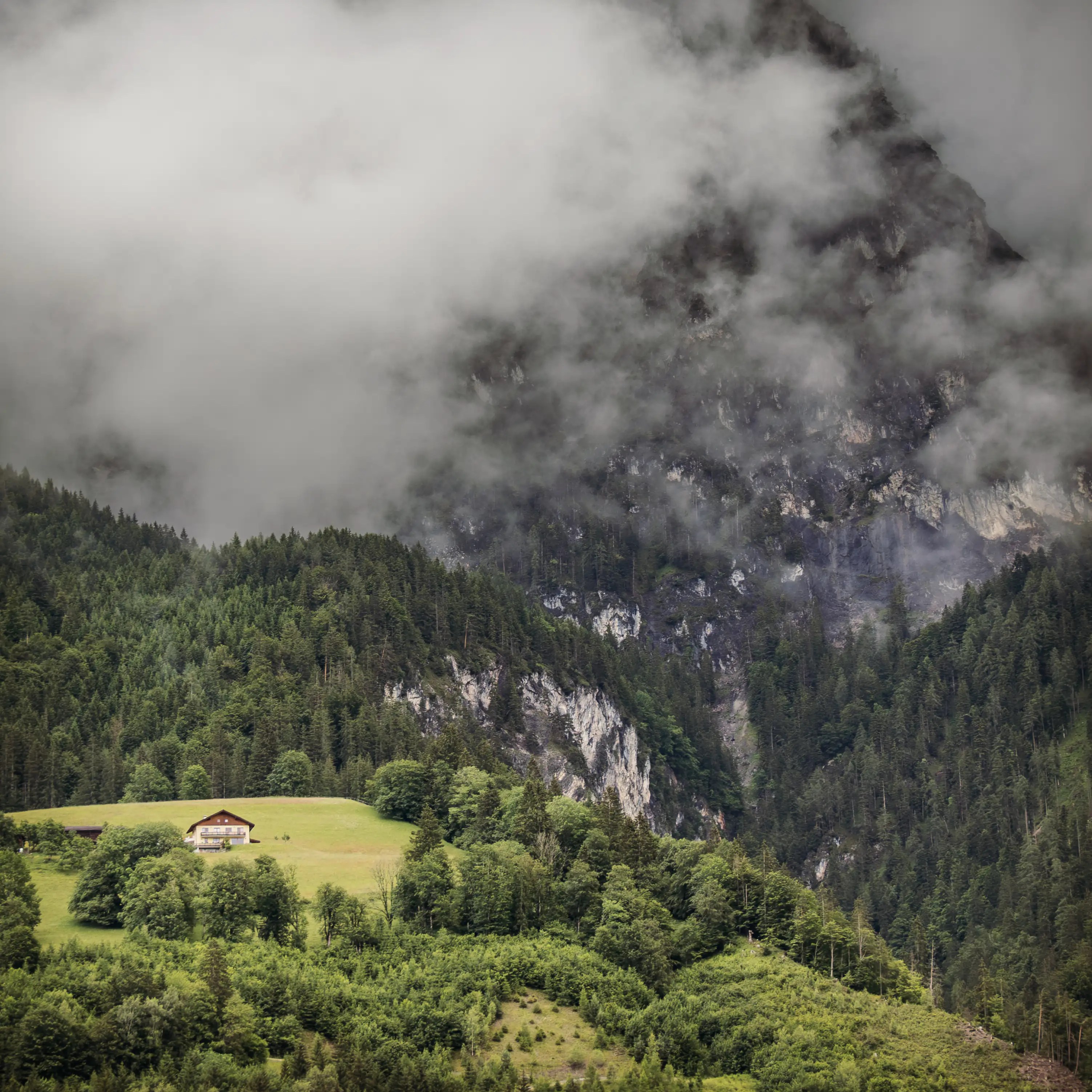 Low clouds drift across steep, forested mountains rising above green fields near the valley below Burg Hohenwerfen.