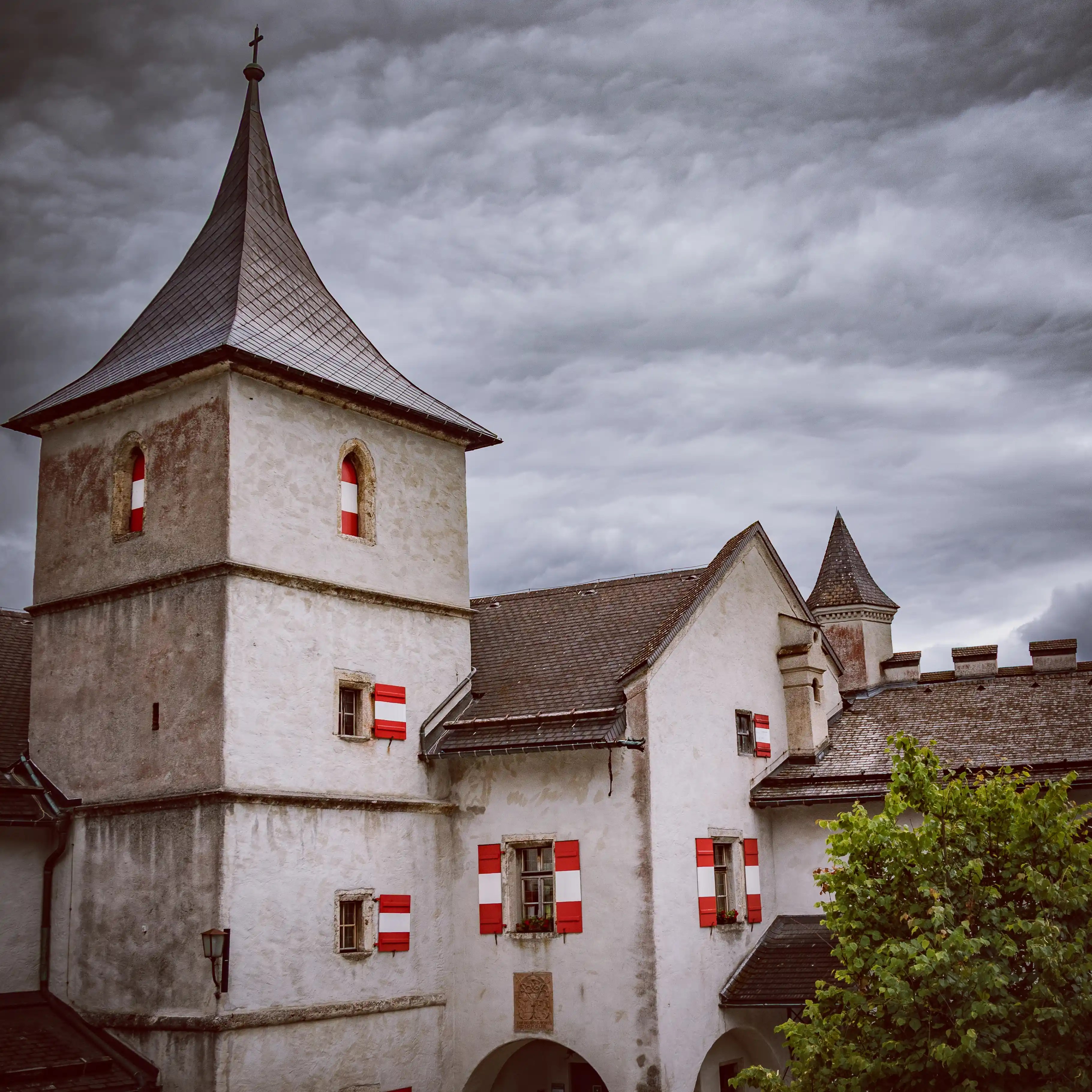 Gray castle buildings with red-and-white shutters and a small tower stand beneath a cloudy sky at Burg Hohenwerfen.
