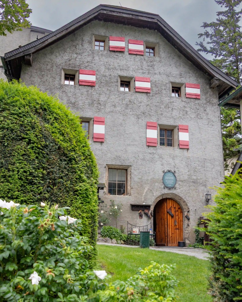 A stone building with red-and-white shutters stands inside the grounds of Burg Hohenwerfen, framed by hedges and greenery.