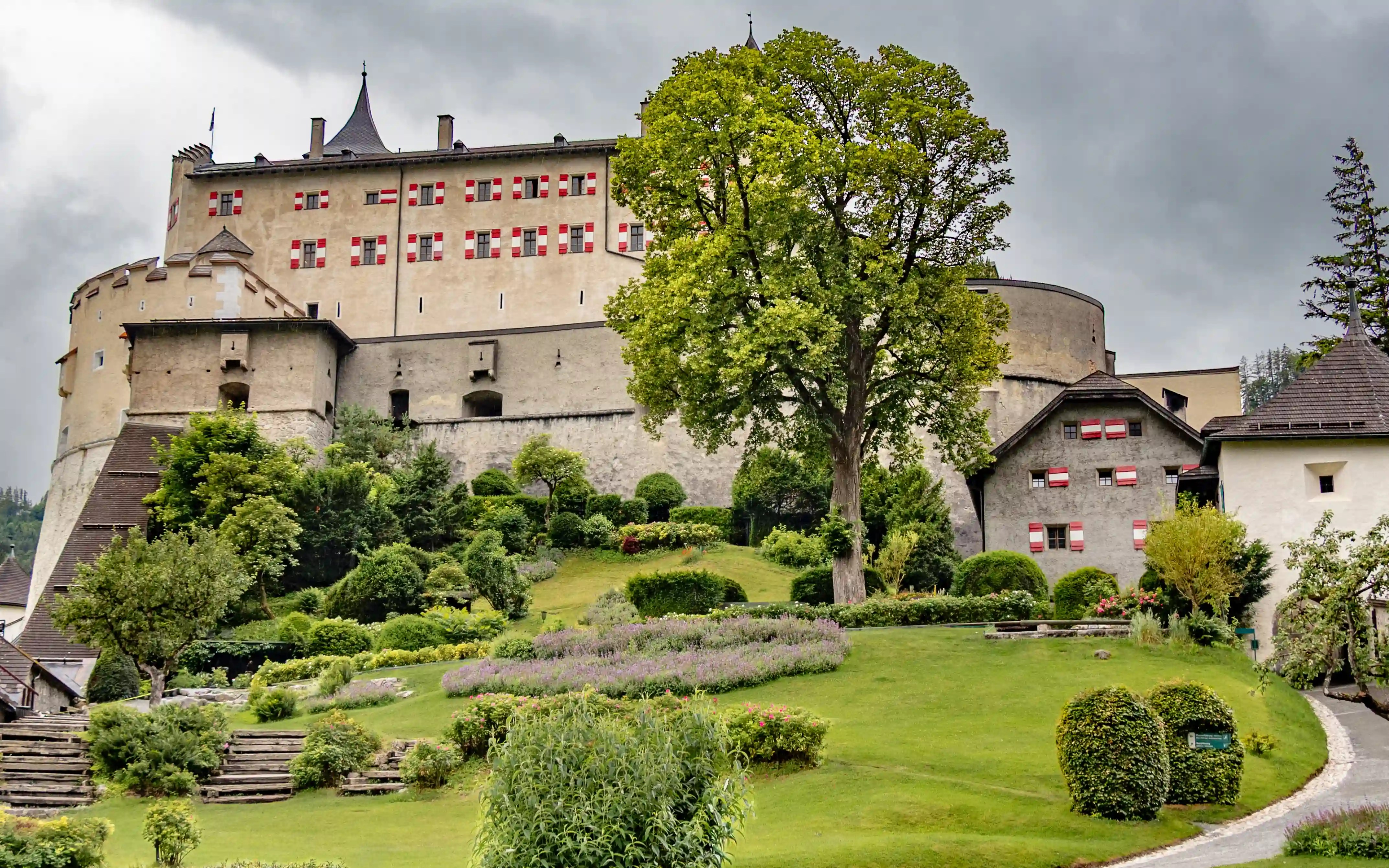 A wide view of Burg Hohenwerfen shows the fortress walls above landscaped gardens and paths on a hillside.