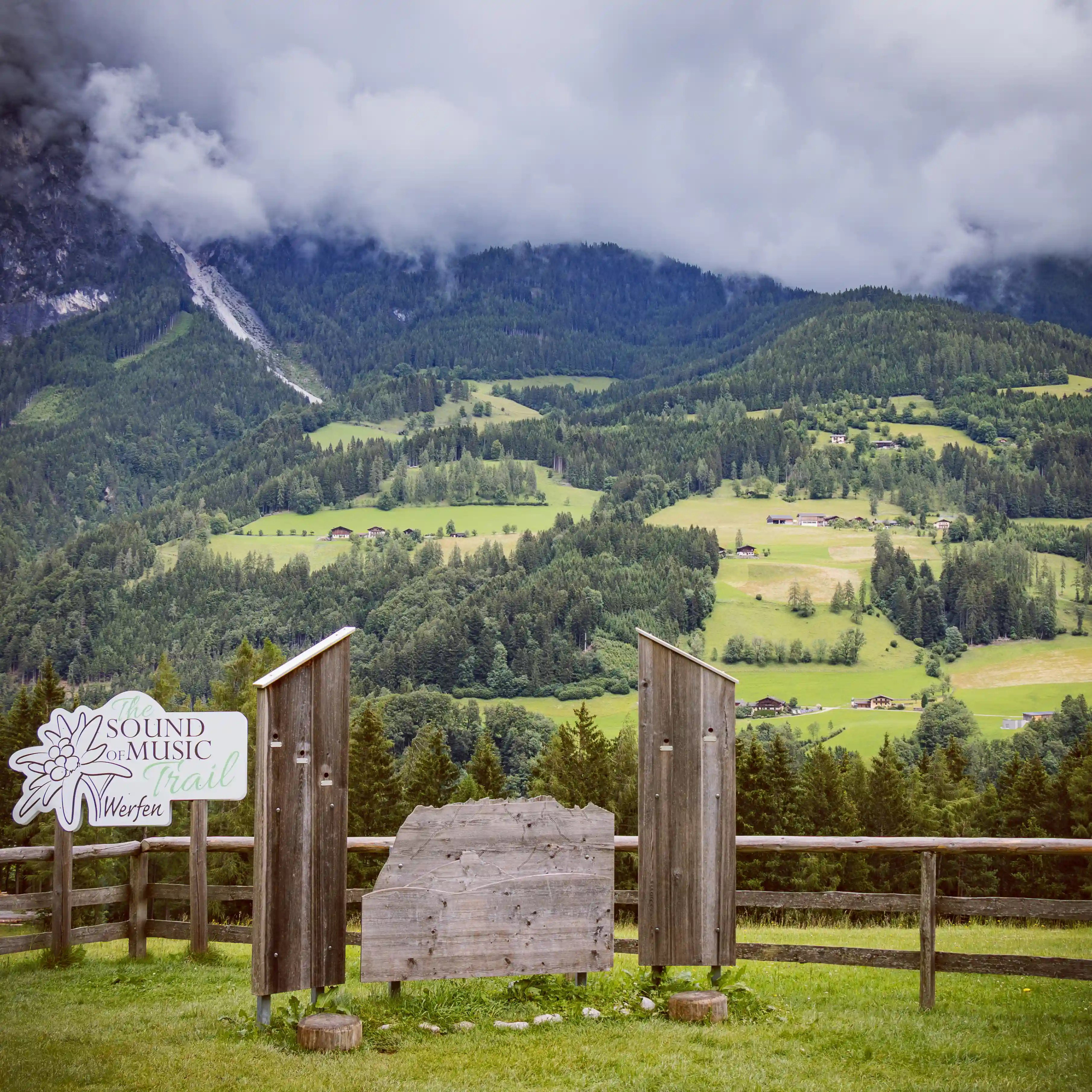 A wooden display along the Sound of Music Trail in Werfen overlooks a green Alpine valley with scattered farmhouses and cloud-covered mountains in the distance.