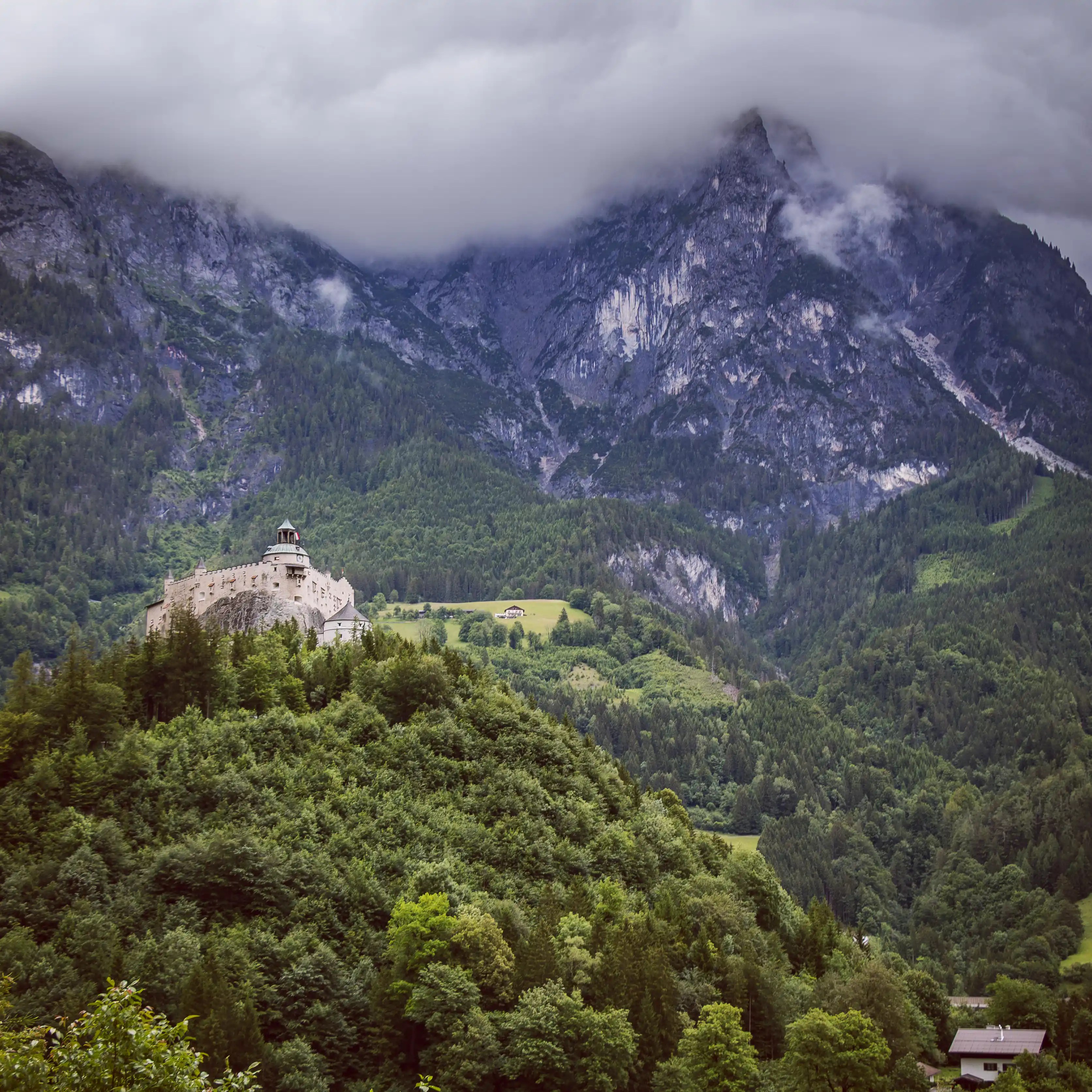 Burg Hohenwerfen sits atop a wooded hill, backed by steep rocky mountains partially hidden by clouds.