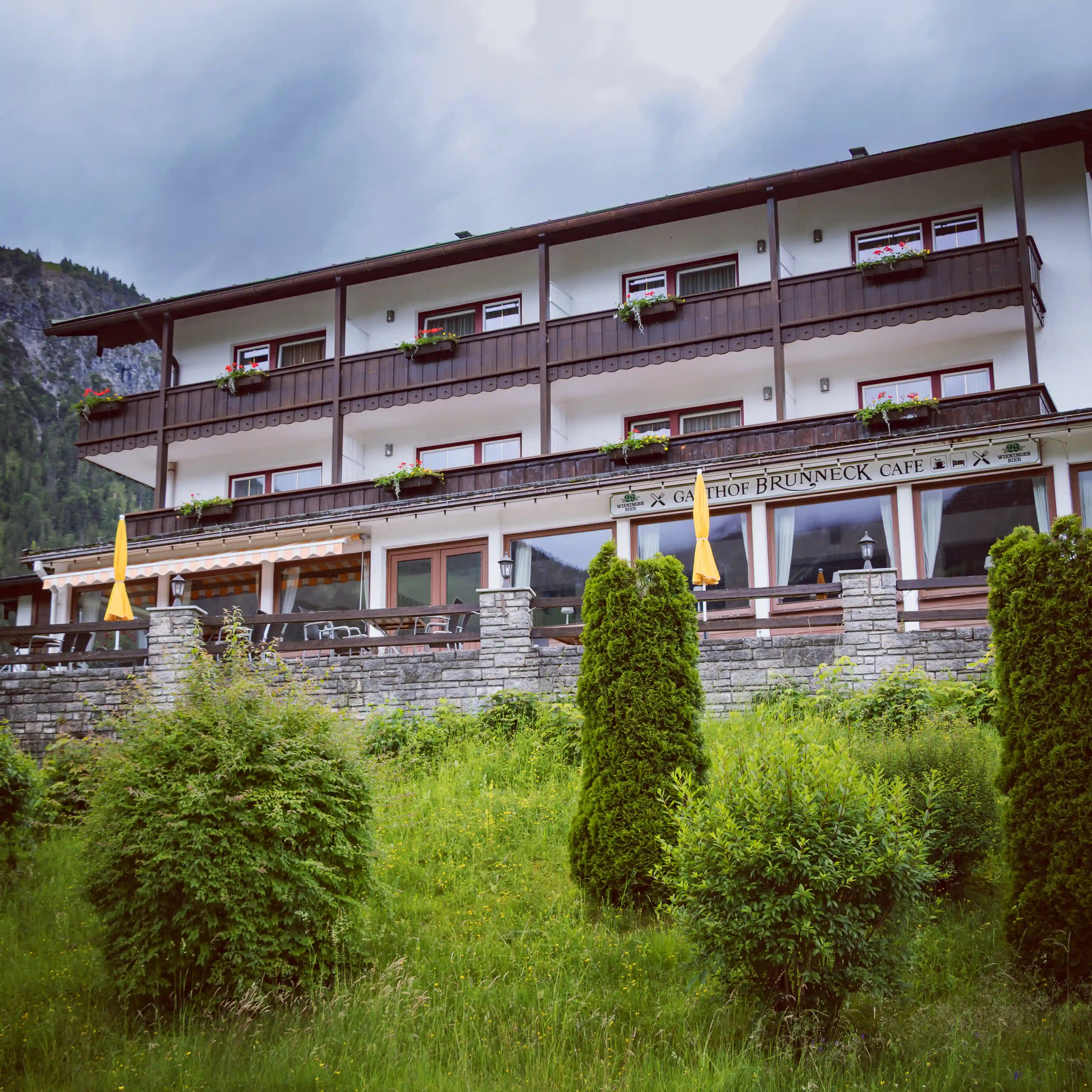 A multi-story alpine-style building with wooden balconies, flower boxes, and a terrace labeled Brunneck Café, set against green hills near Königssee.